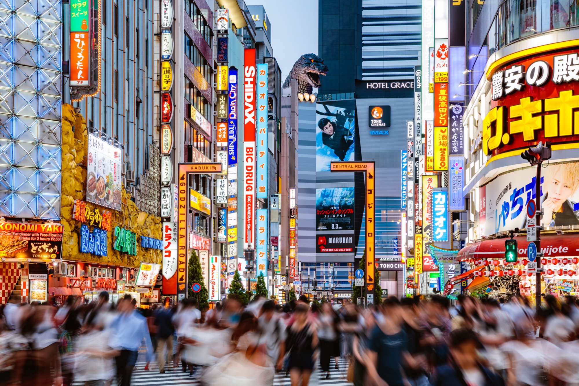 Belebte Straße in Tokios Stadtteil Kabukicho mit leuchtenden Reklametafeln, Menschenmengen und Godzilla-Kopf auf einem Gebäude.
