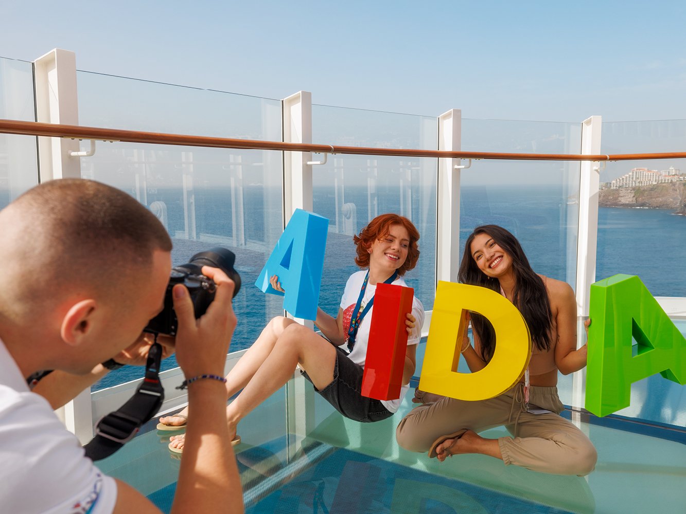 Ein Fotograf macht ein Foto von zwei Teenagern, die an Deck sitzen und große bunte AIDA Buchstaben halten
