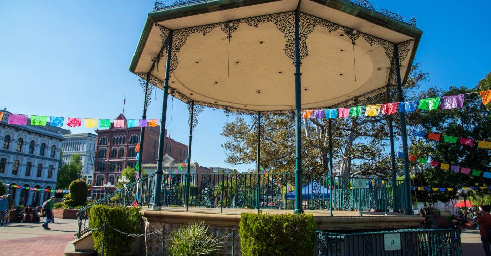 a round green pergola in the middle of a courtyard surrounded by people, lush green trees and buildings at El Pueblo de Los Angeles Historical Monument with blue sky in Los Angeles California
