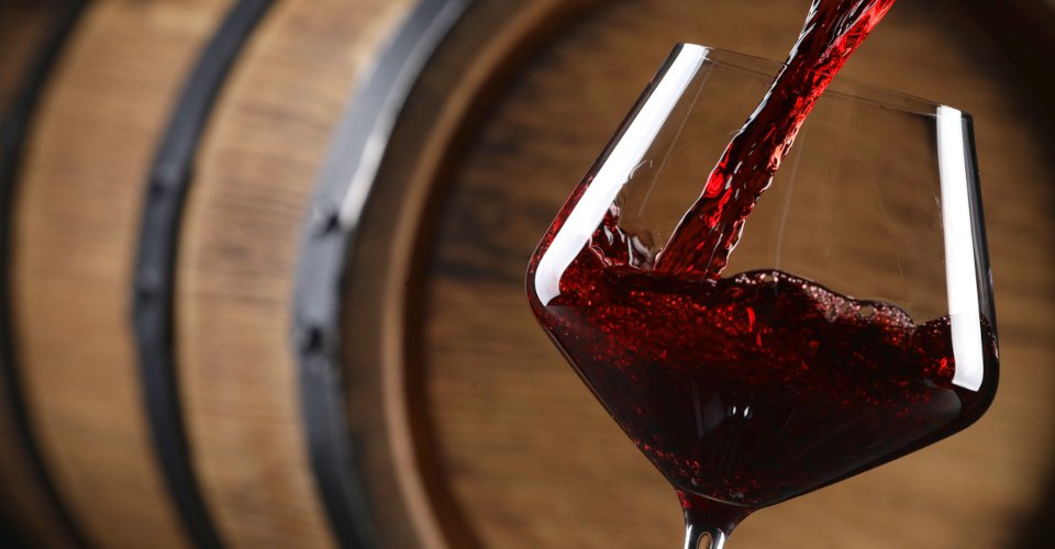Pouring red wine into glass near wooden barrel, closeup. Space for text