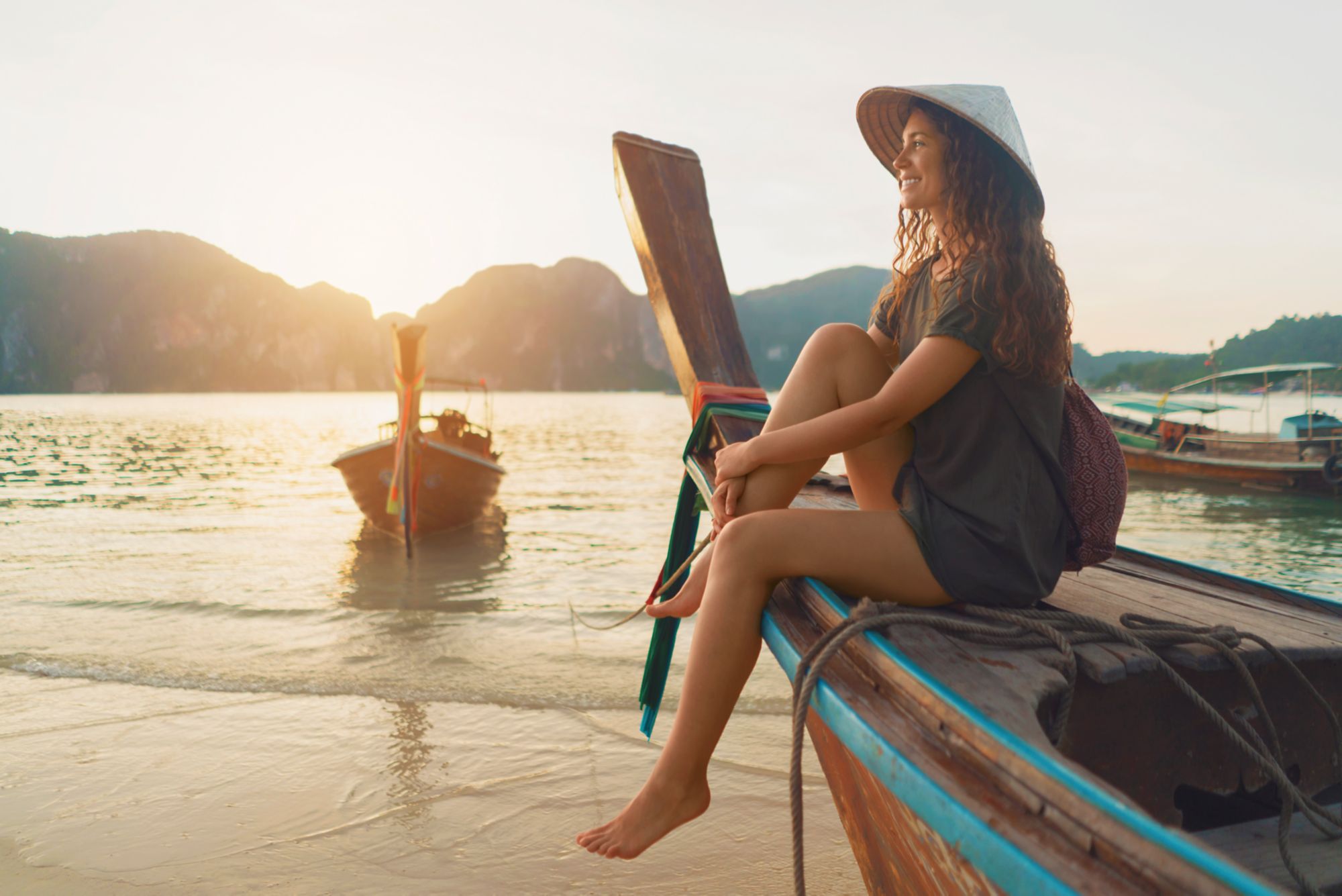 Schöne kaukasische Frau mit asiatischem, konischem Hut sitzt auf einem Longtail-Boot und genießt einen unbeschwerten Sommertag am Strand.