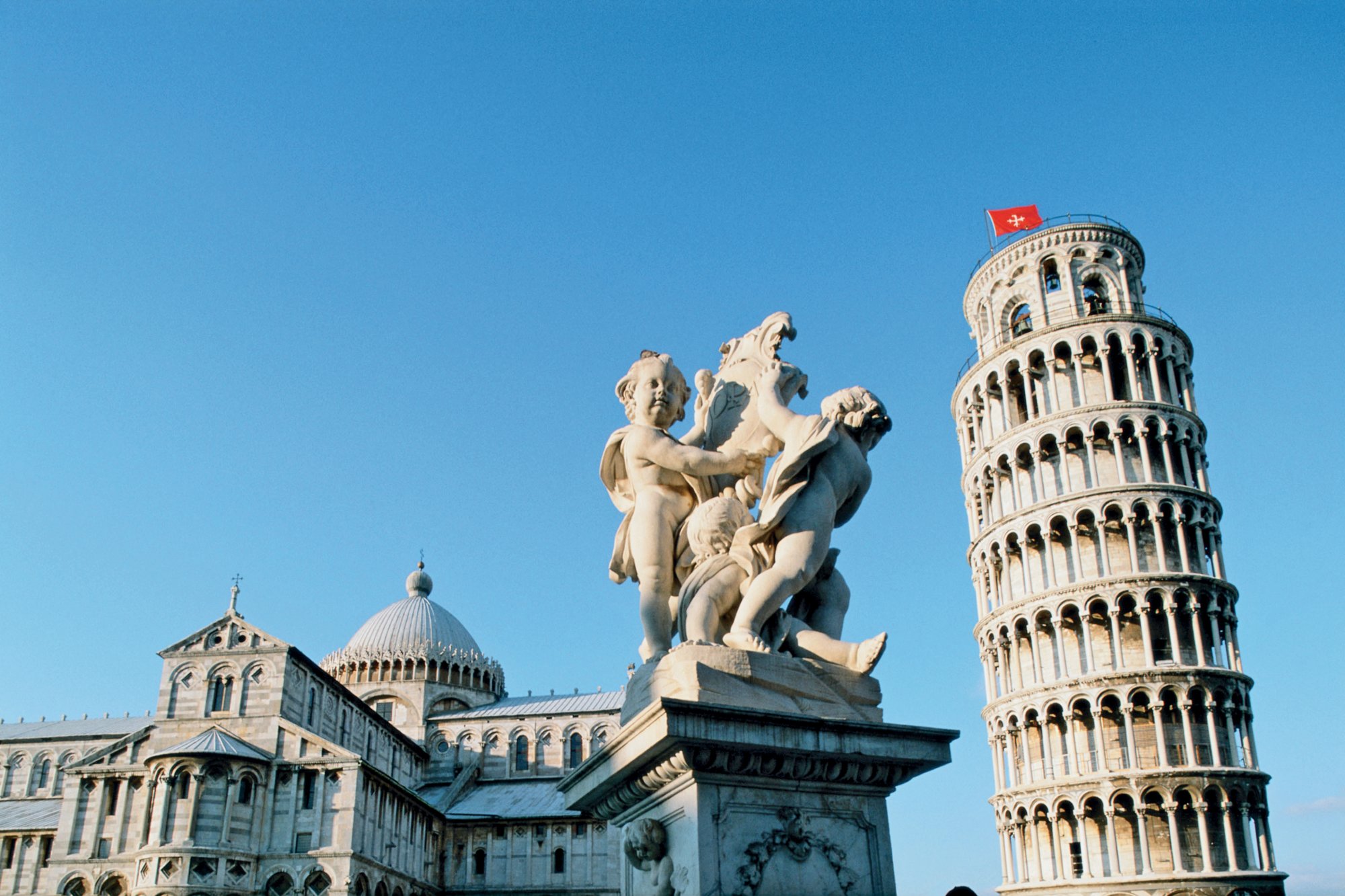 Blick auf den Schiefen Turm von Pisa mit Marmorskulptur im Vordergrund und strahlend blauem Himmel.