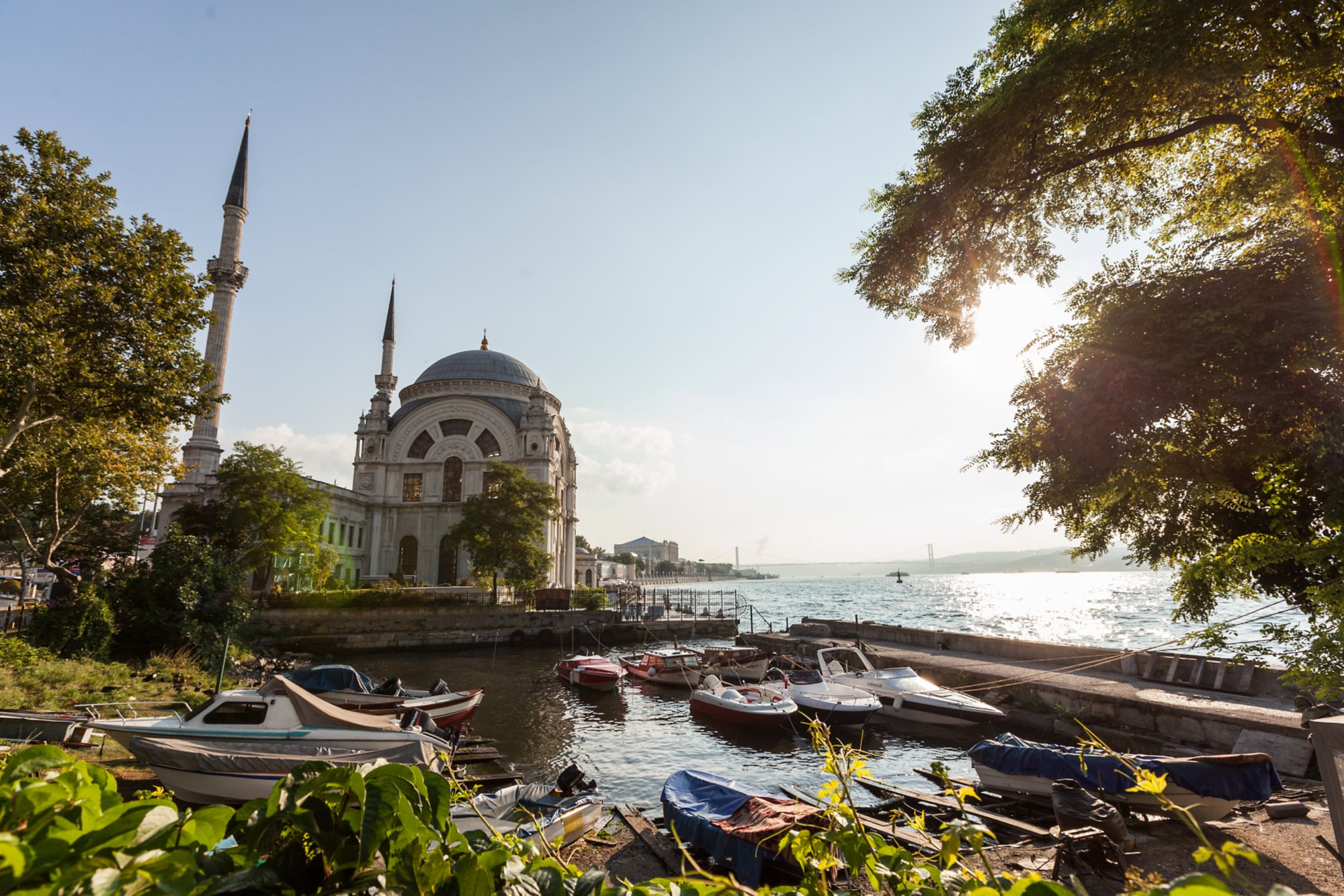 Moschee mit zwei Minaretten am Wasser in Istanbul, umgeben von Booten, Sonnenlicht und städtischer Kulisse im Hintergrund.