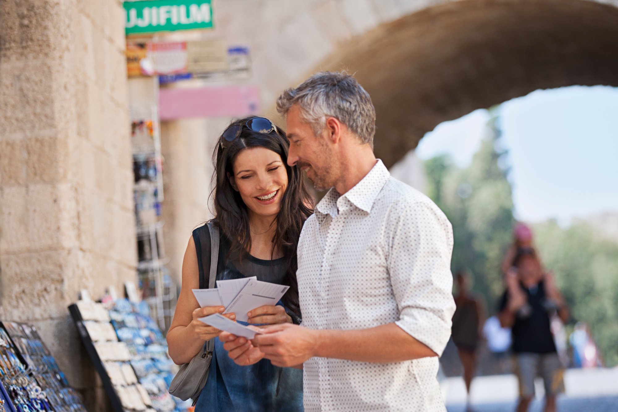 Touristen in Griechenland betrachten Postkarten vor historischem Steinbogen und Souvenirstand.