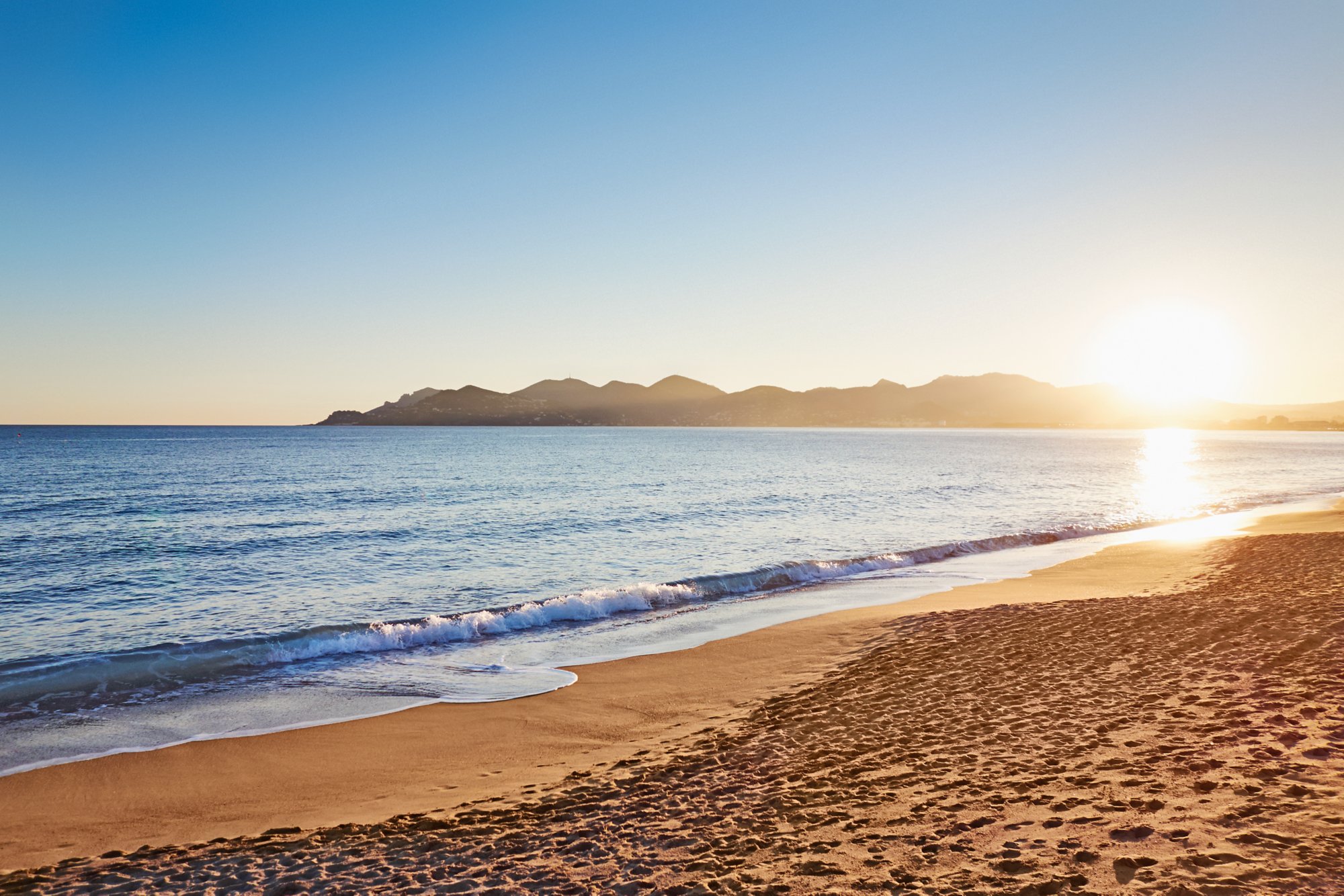Rechts unten ist der goldene Sand, das Meer spült in sanften Wellen über den Sand und läuft links mittig aus dem Bild. Im Hintergrund verläuft eine felsige Küste aus dem rechten Bildrand raus. Die Sonen geht auf der rechten Seite gerade über den Felsen auf.