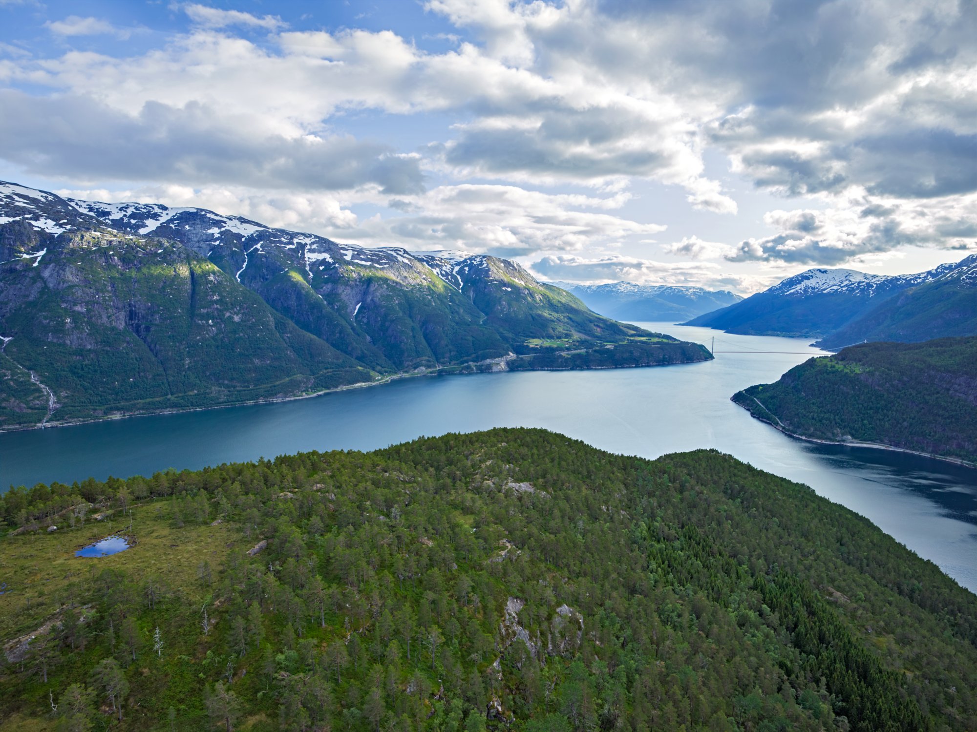 Blick auf den ruhigen Eidfjord, umgeben von schneebedeckten Bergen und grüner Vegetation unter teils bewölktem Himmel.