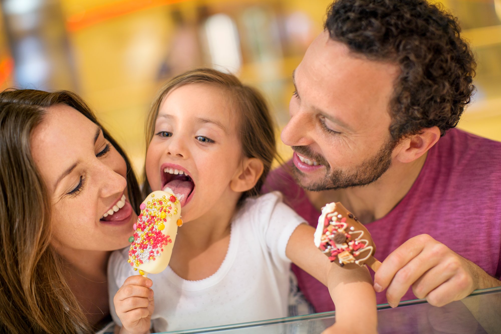 Eine dreiköpfige Familie isst zusammen ein Eis. Das kleine Mädchen und der Vater haben ein reich verziertes Eis in der Hand. Das Mädchen und die Mutter wollen von dem Eis des Mädchens abbeißen. Alle haben braune Haare, der Vater trägt ein beerefarbendes T-Shirt und das Mädchen ein weißes.