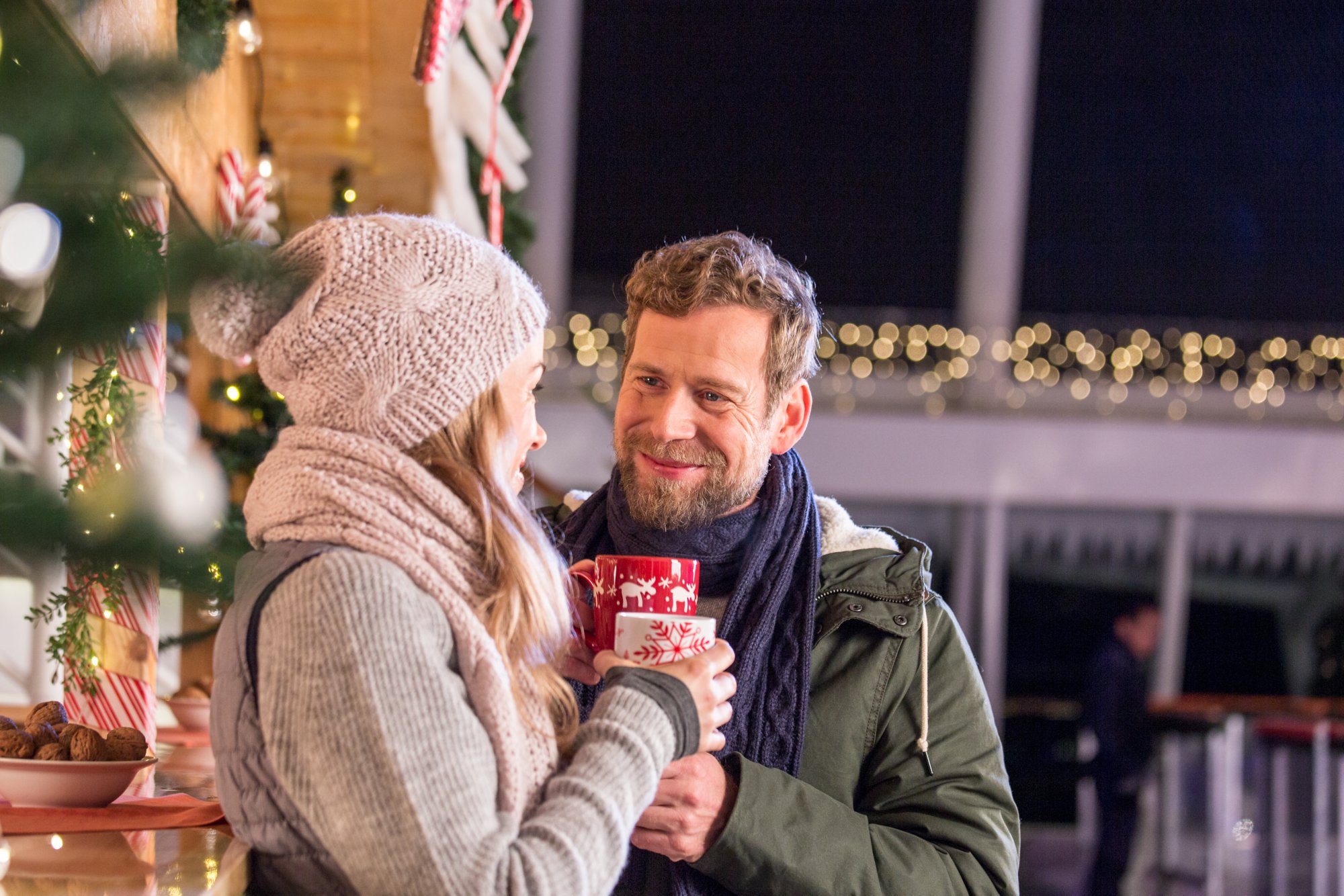 Ein Pärchen steht warm angezogen an einer weihnachtlichen Hütte mit Tassen in der Hand