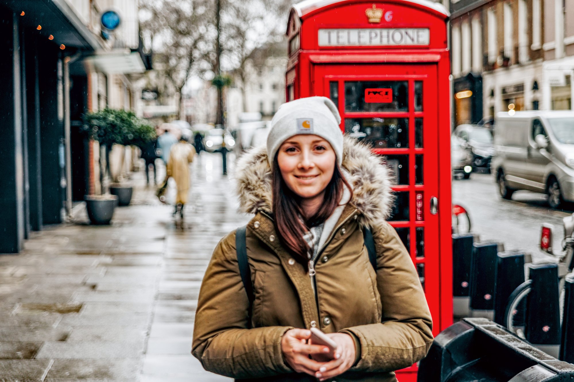 Junge Frau mit warmer Jacke und Mütze bekleidet steht in London vor einer Typischen Telefonzelle