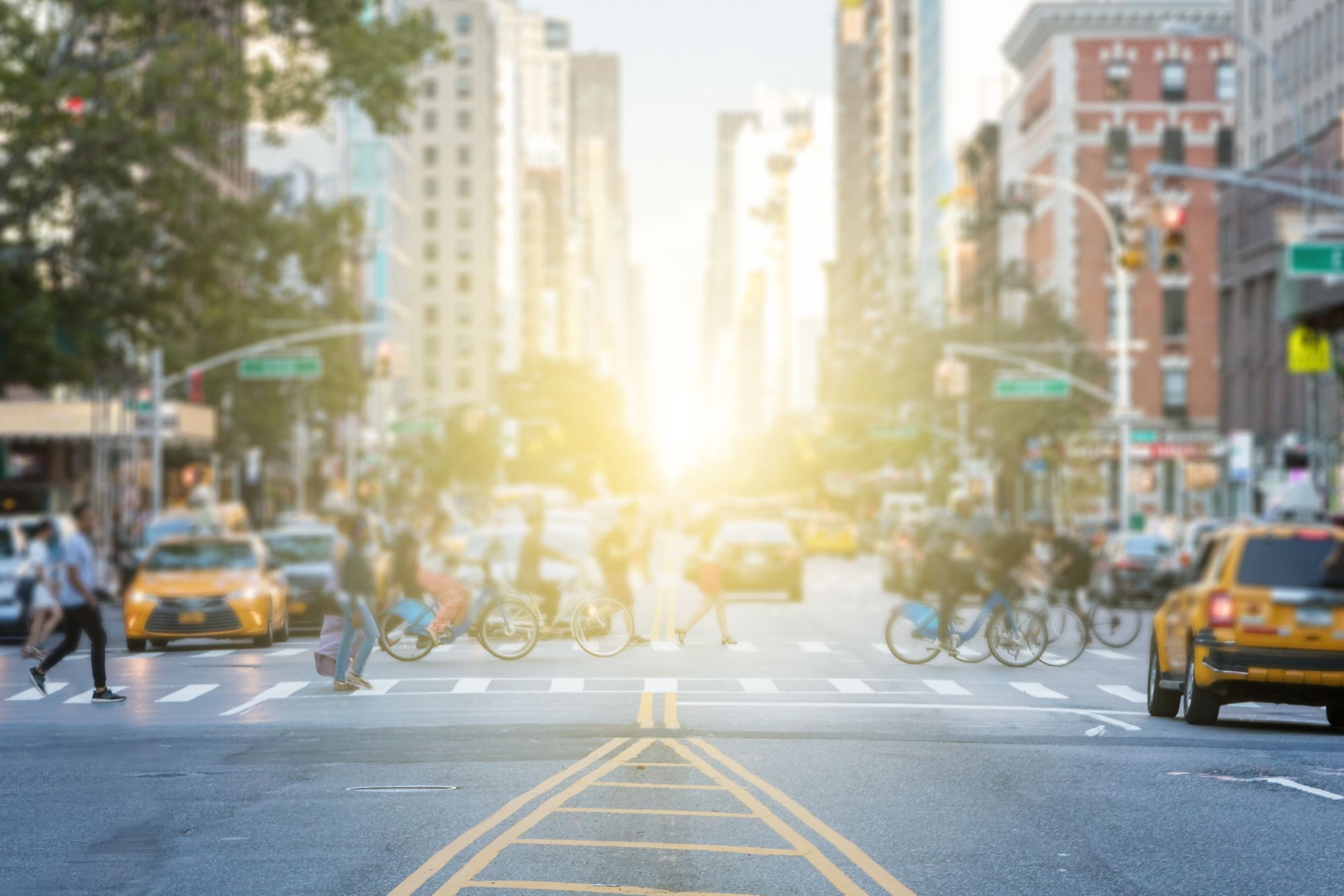 People crossing the busy intersection between traffic on 3rd Avenue and 10th Street in Manhattan in New York City with the glow of sun light in the background