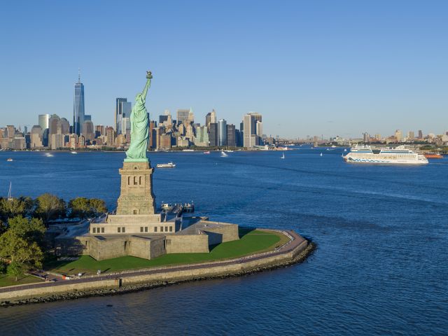Freiheitsstatue vor der Skyline von Manhattan, dahinter ein Kreuzfahrtschiff im Hudson 