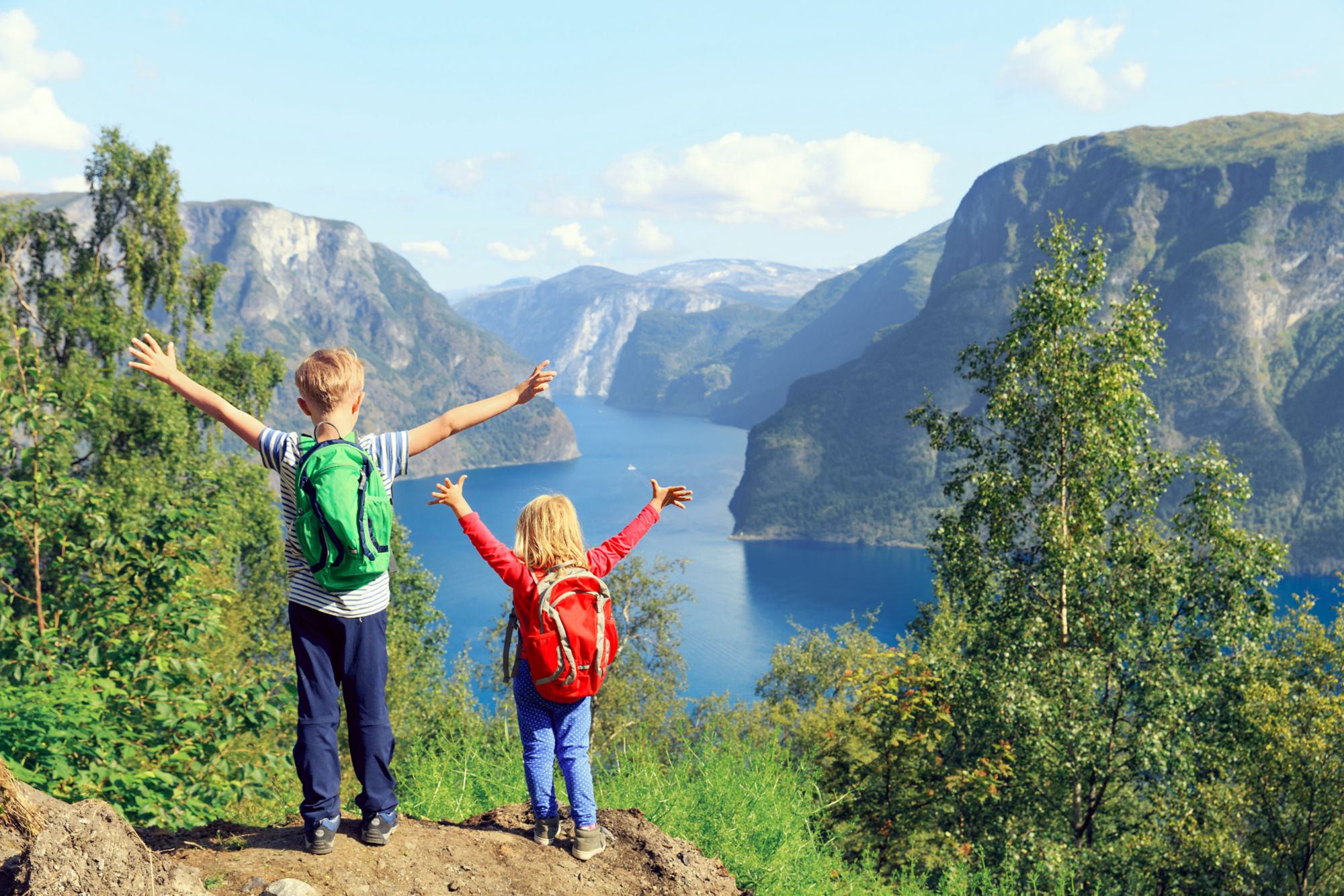 Zwei Kinder mit einem Rucksack stehen oberhalb eines Fjordes 