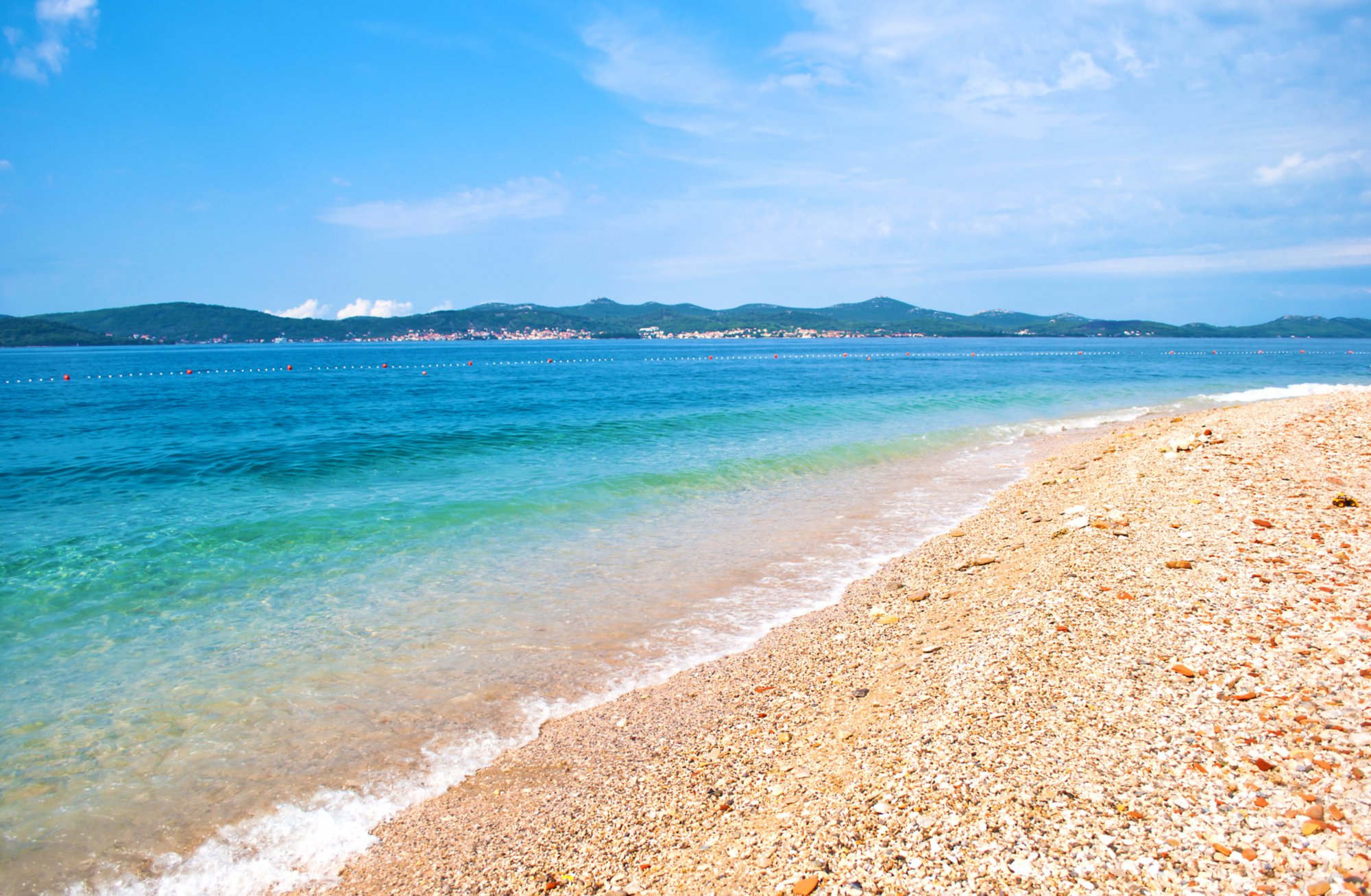 Pebble sandy beach on the background of a hill range with many small houses, a line of buoys and azure transparent blue sea. Blue cloudless sky. Zadar, Croatia