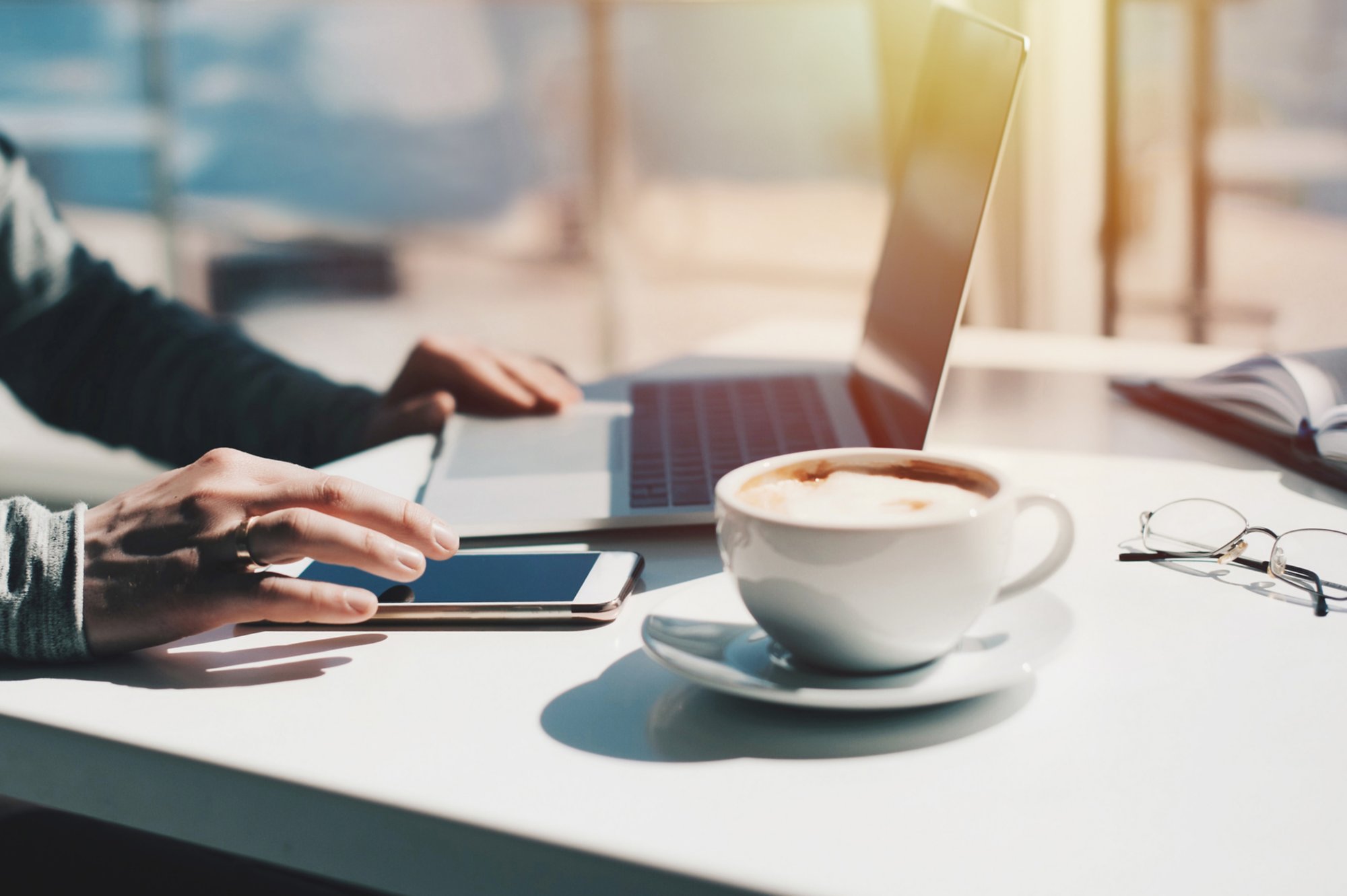 Ein Laptop steht aufgeklappt auf einem Tisch, die Hände eines Mannes sind zu sehen. Neben dem Laptop liegt ein Mobiltelefon und eine Tasse Kaffee