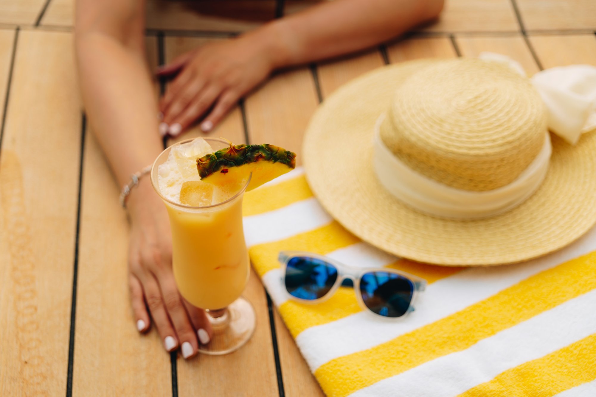 Gelb-weiß gestreiftes Handtuch, darauf liegen Sonnenhut und Sonnenbrille, eine Frau hält daneben ein Cocktailglas