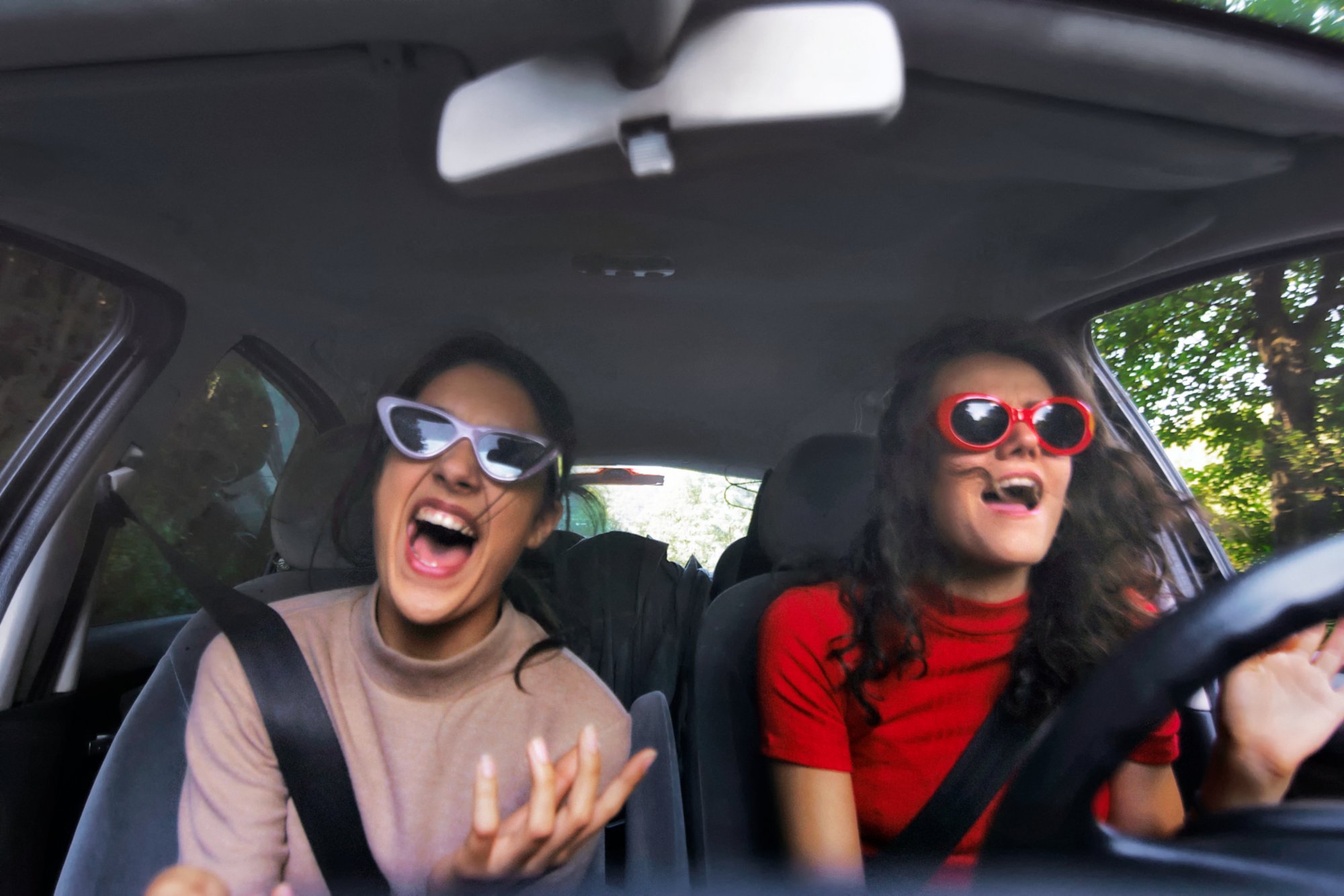 Zwei junge Frauen im Auto singen ausgelassen zu einem Lied Radio. Sie haben Sonnenbrillen auf und die linke trägt einen pastell-rosa farbenden Pulli und die anderen einen knallroten