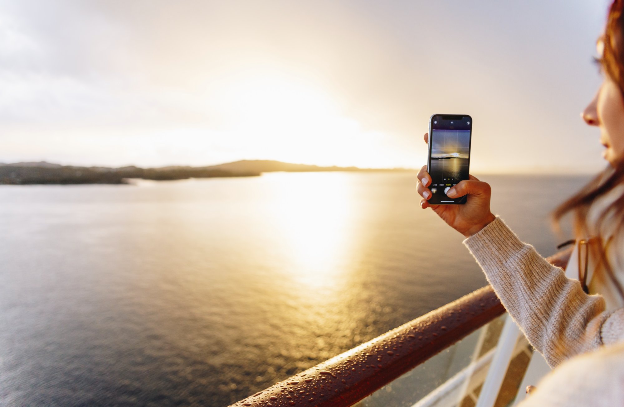 Eine Frau steht an Deck und fotografiert den Sonnenuntergang mit einem Smartphone