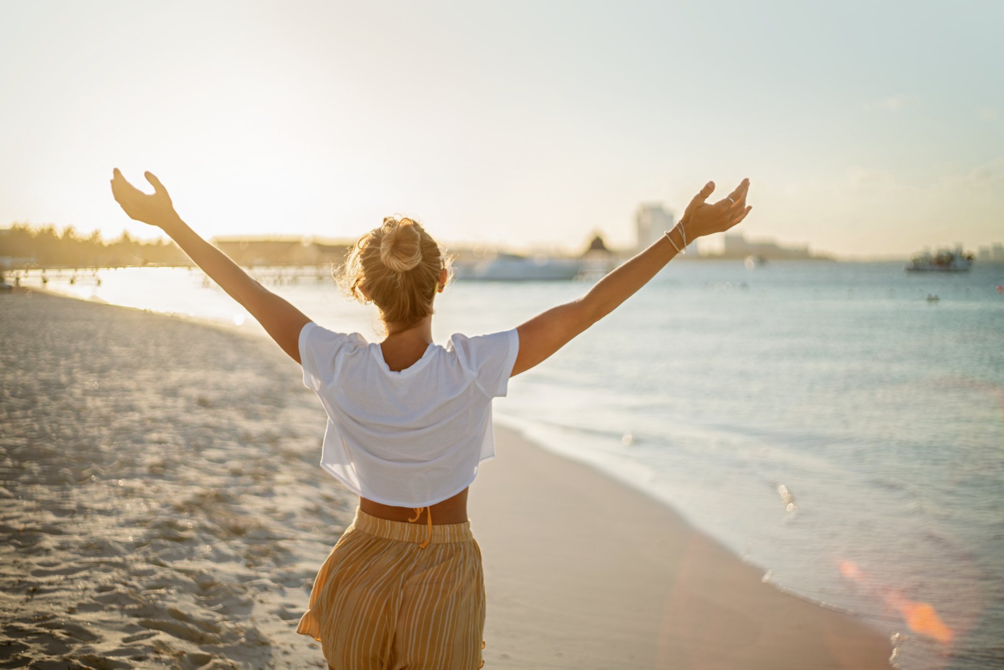 Cheerful young woman embracing nature at sunset; female standing on beach arms outstretched, Cancun, Mexico