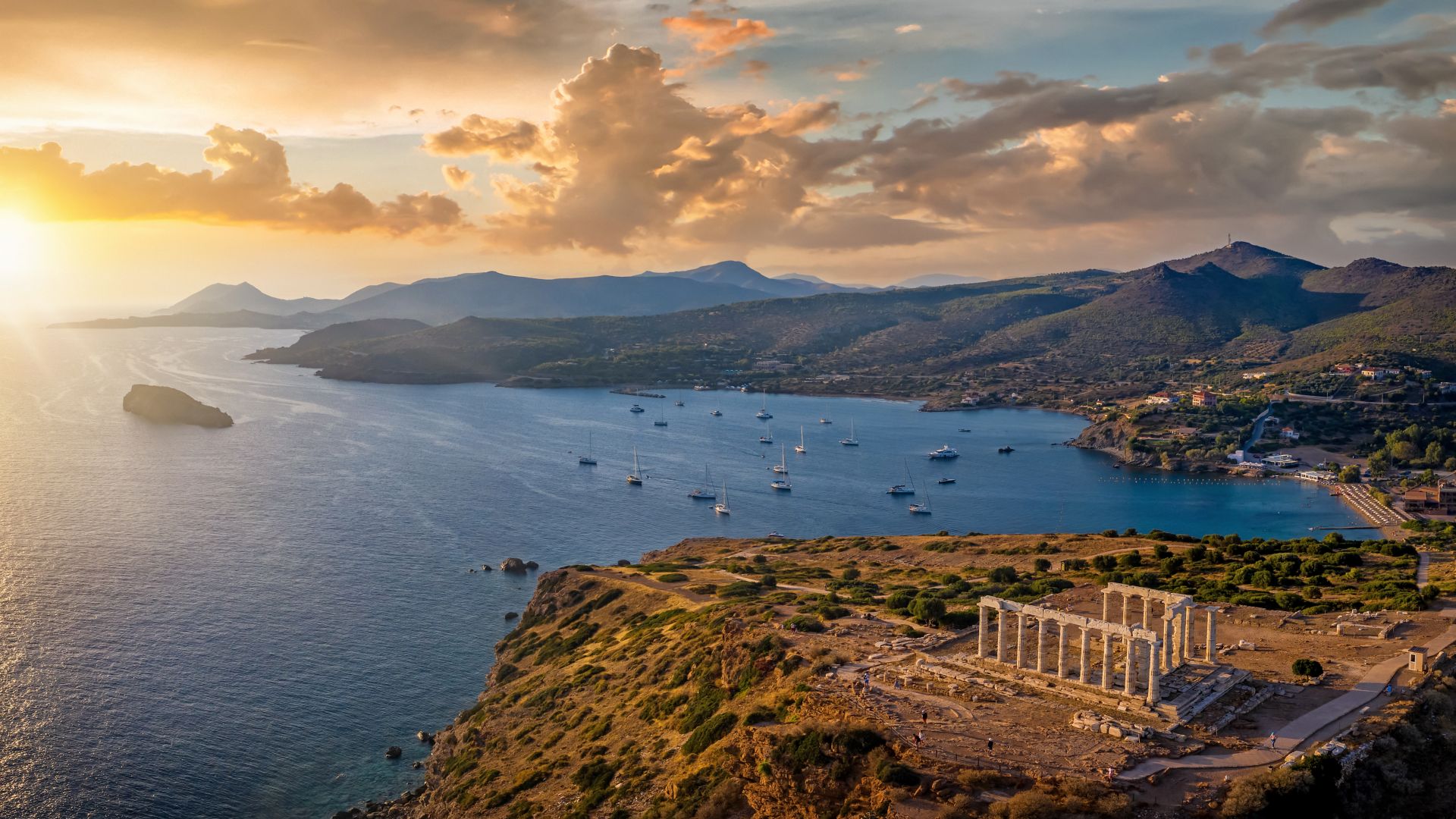 Panoramic view of the Temple of Poseidon at Cape Sounion at the edge of Attica, Greece, with moored sailboats in the bay during sunset time