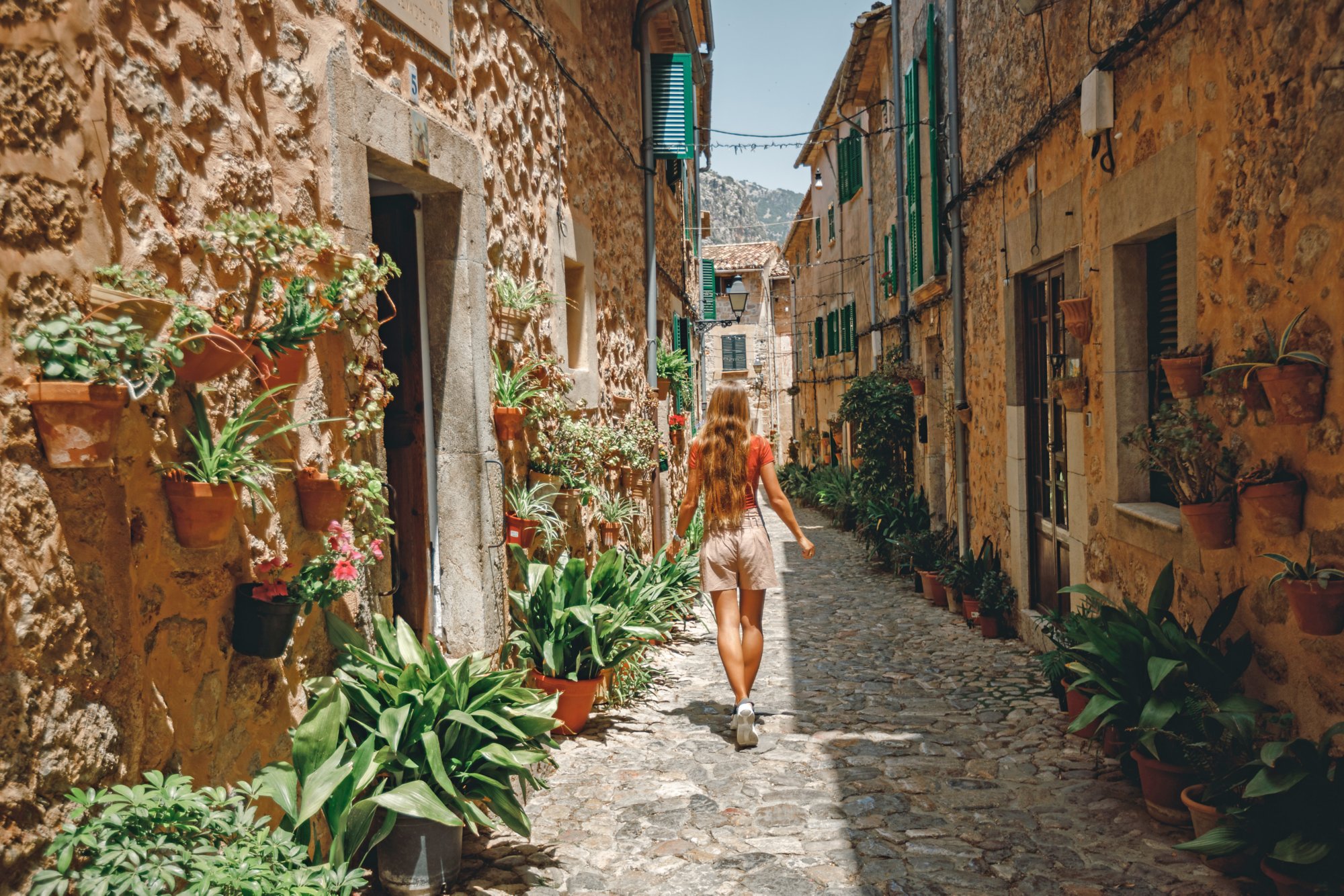 Back view young girl tourist walk through cozy and beautiful  street decorated with flower pots and ceramic tiles in Valldemossa, Mallorca, Spain. Vacation in Spain