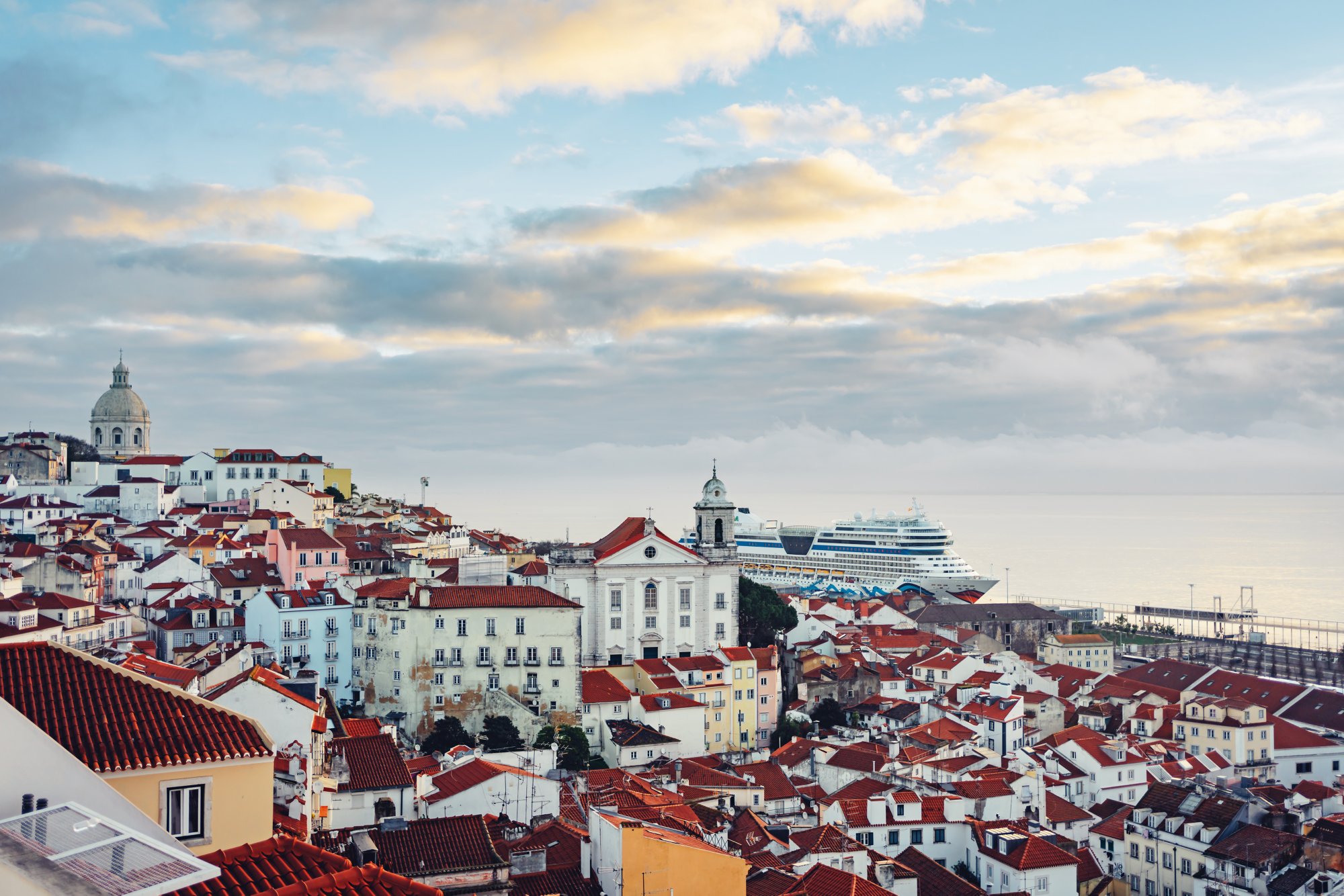 Panoramablick auf Lissabon mit roten Ziegeldächern, weißer Kirche und AIDA Kreuzfahrtschiff im Hafen bei sanftem Licht.
