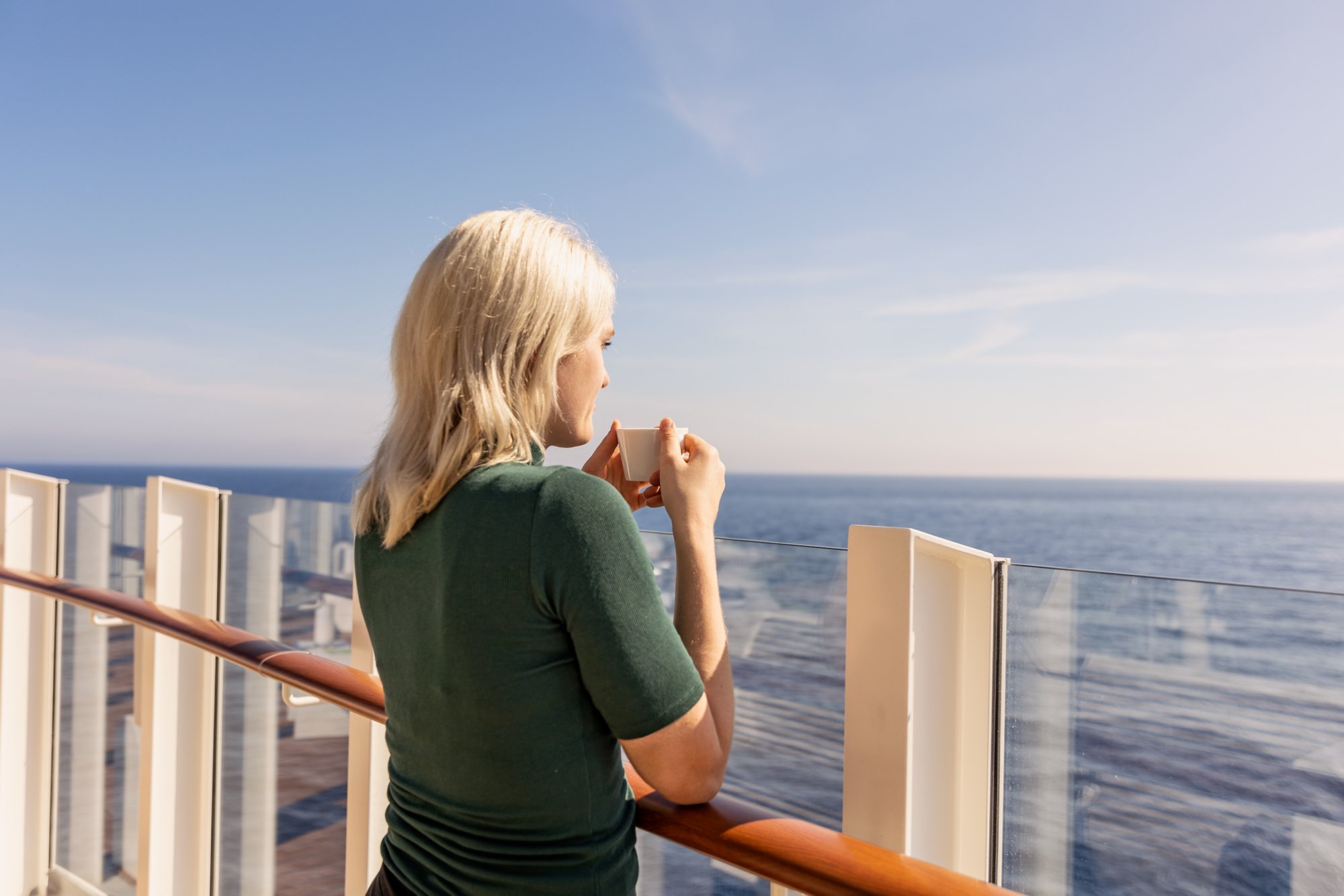 Eine Frau steht an der Reling an Deck, hält eine Tasse und blickt auf das Meer