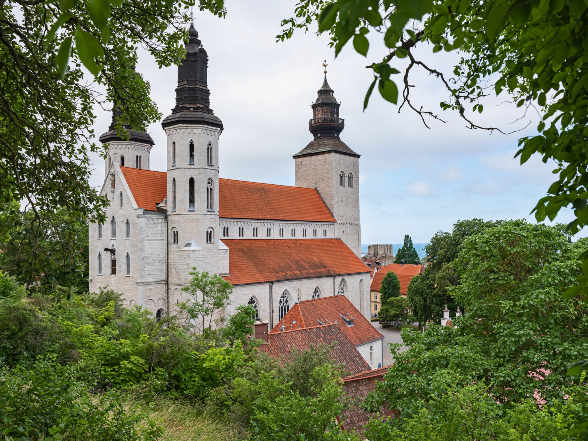 Historische Kirche mit drei Türmen in Visby, Schweden, umgeben von grüner Vegetation und roten Ziegeldächern.
