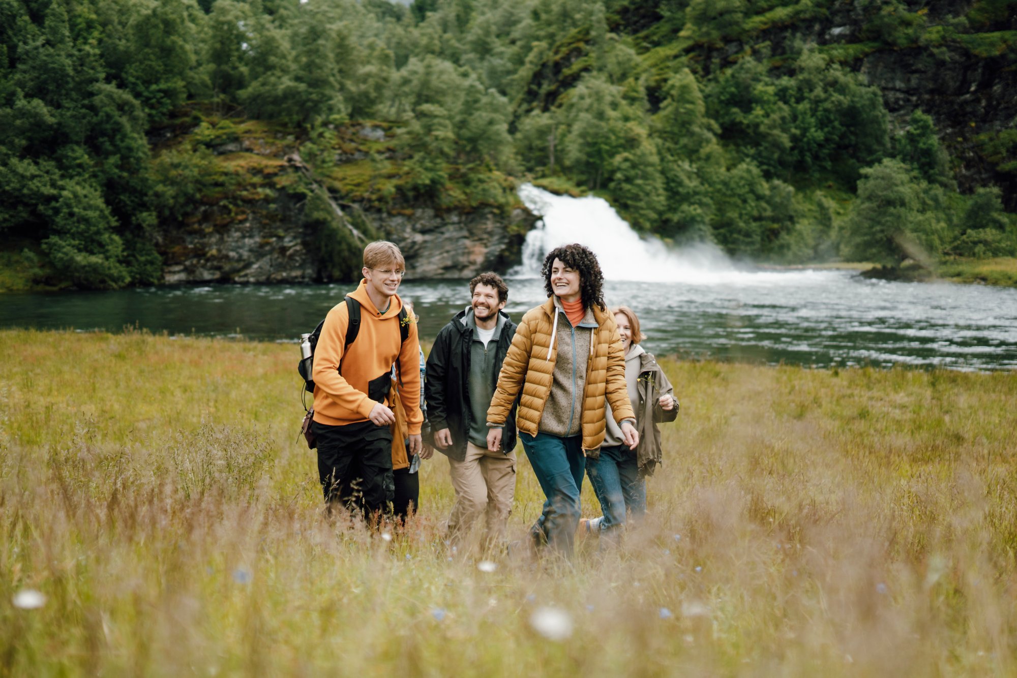 Gruppe wandert über Wiese vor einem Wasserfall und Wald