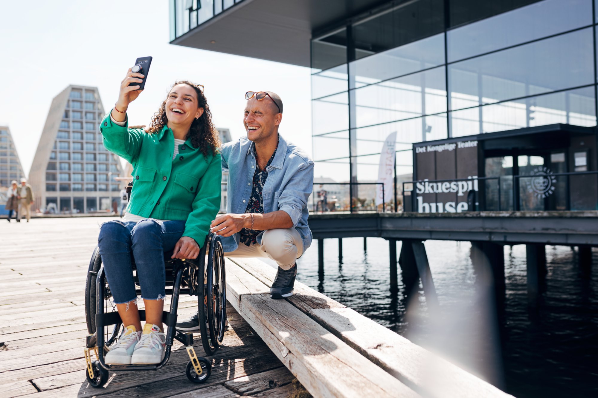 Eine junge Frau sitzt im Rollstuhl, ein Mann hockt dahinter, sie macht ein Selfie von den beiden