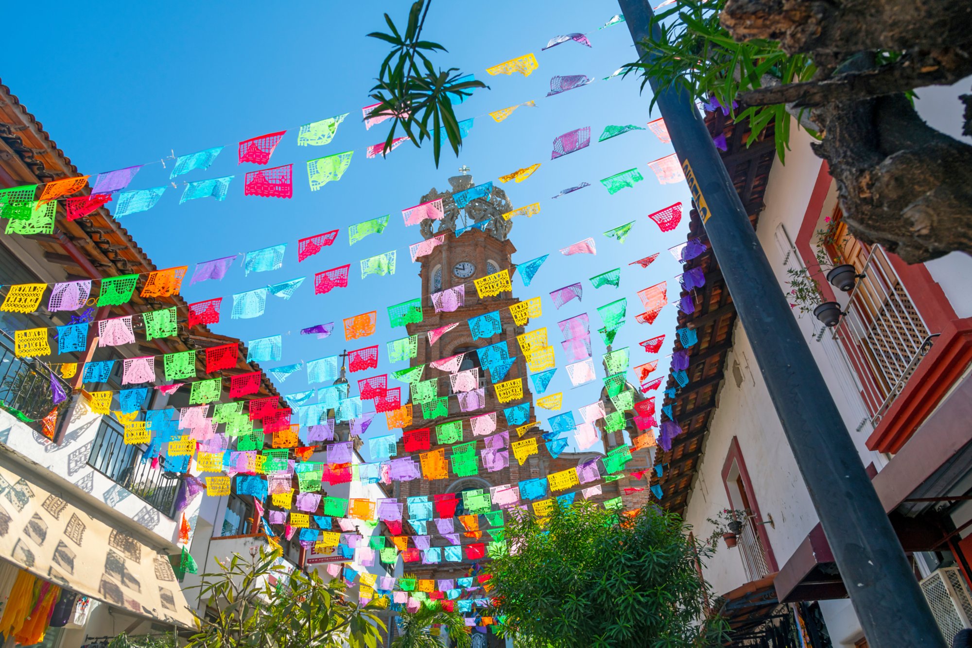 View from the street below of the The Church of Our Lady of Guadalupe with colorful flags strung across the street on a sunny morning in Puerto Vallarta, Mexico.