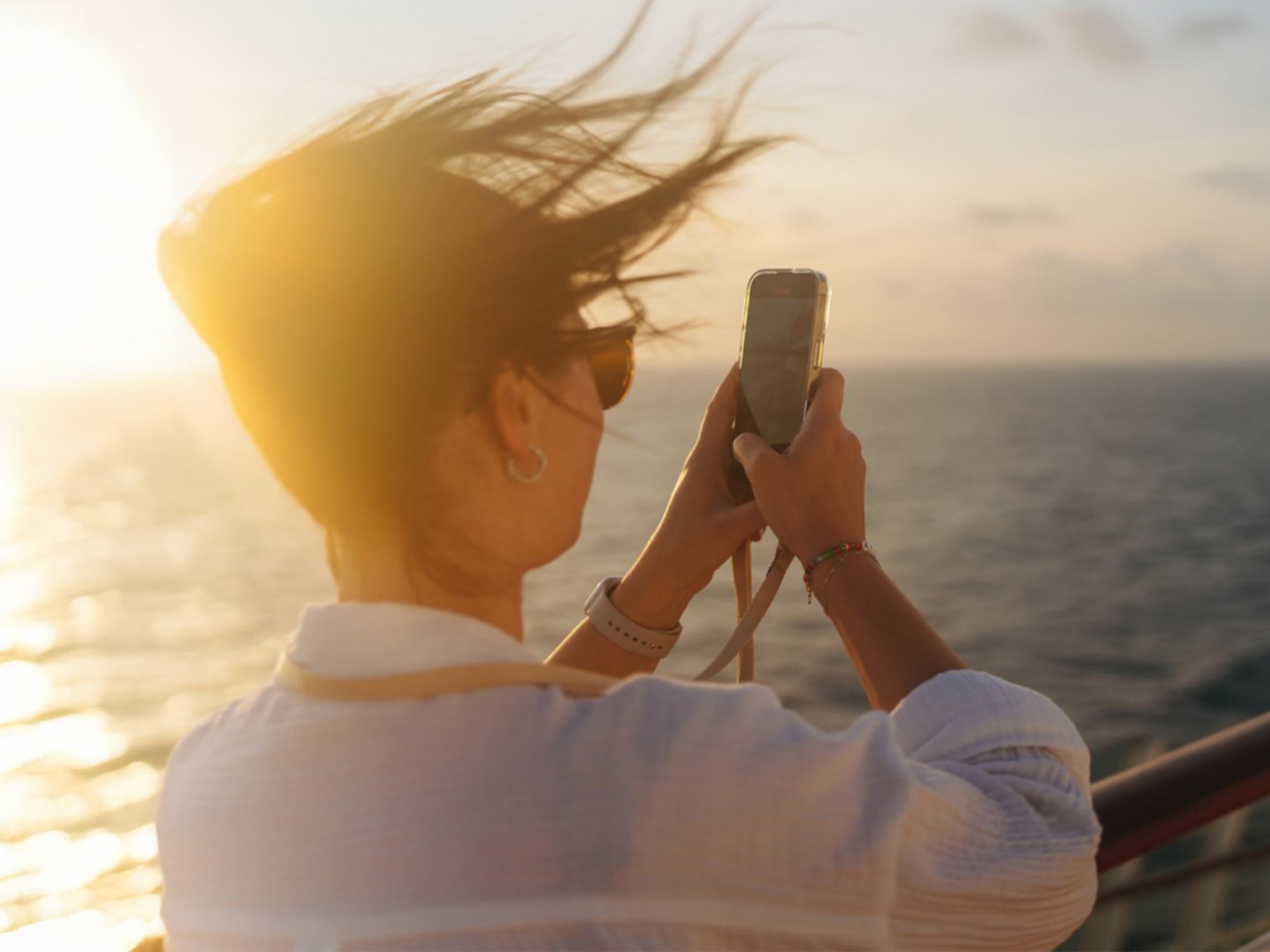 Junge Frau steht an der Reling und macht ein Foto mit ihrem Smartphone vom Sonnenuntergang auf dem Meer
