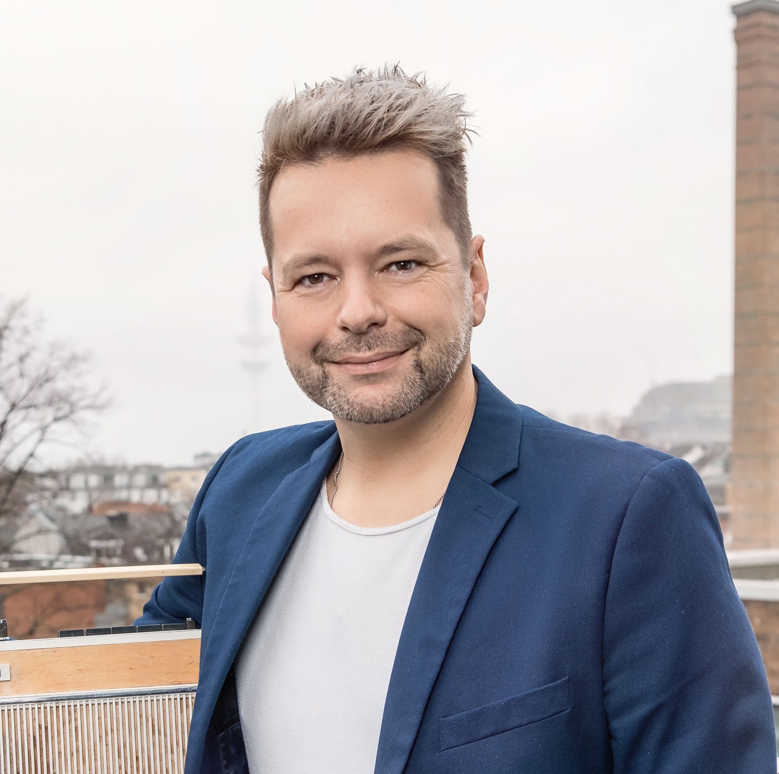 Mann mit verhaltenem Lächeln, Dreitage-Bart und blonden hochgestylten kurzen Haaren. Er trägt eine blaue Anzugjacke und darunter ein weißes T-shirt.