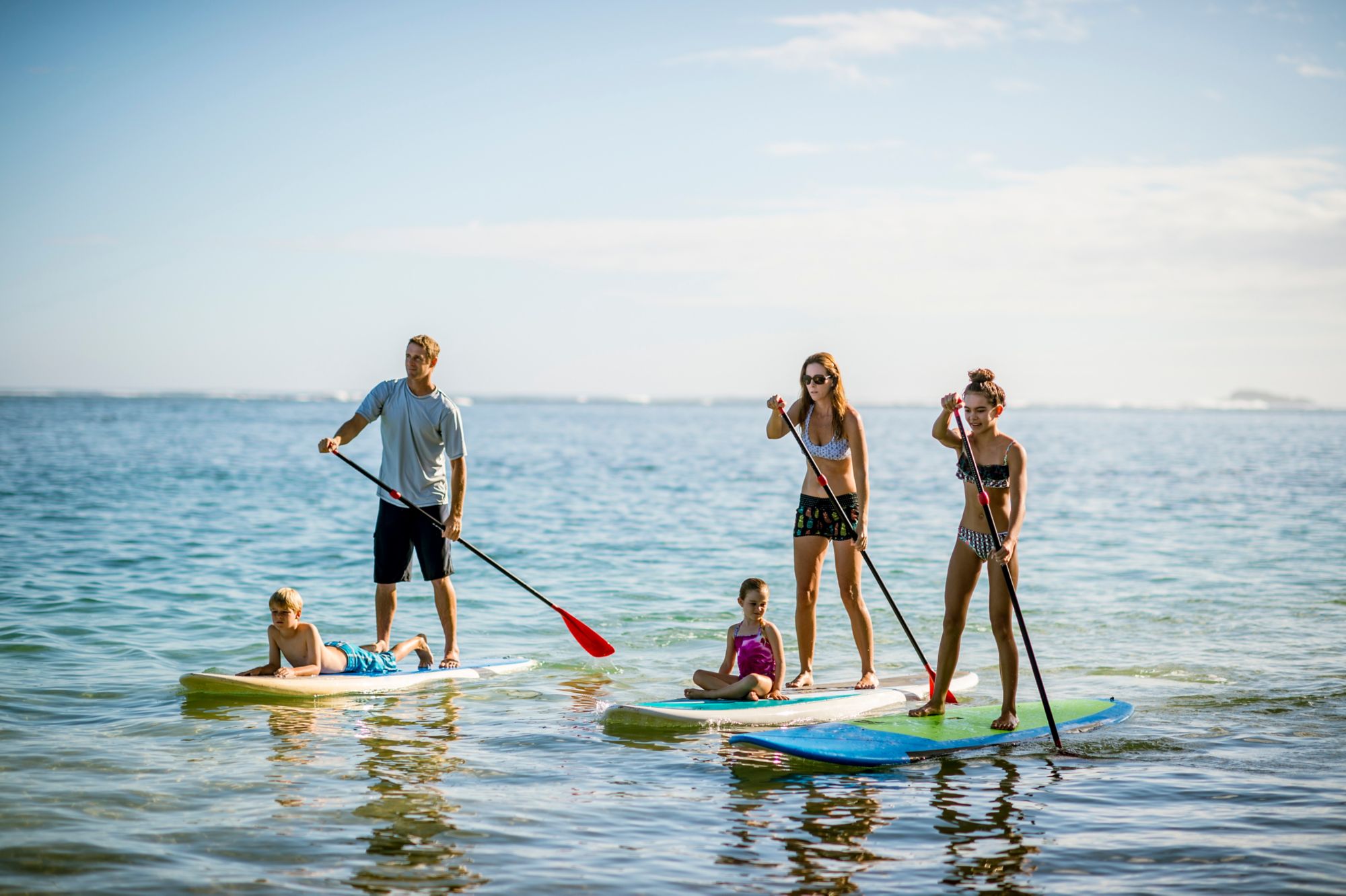 Family in Hawaii's tropical climate enjoying their vacation on stand up paddleboards (SUP) in the sea.