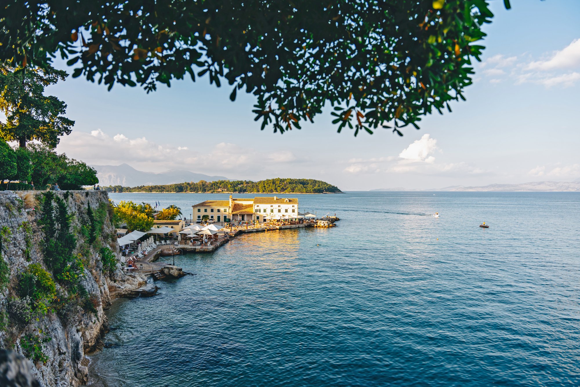 Von links ragt eine seile felsige Küste ins Bild, von dort ein Steg mit Häusern aufs Meer verteilt stehen Sonnenschirme. Das Meer ist blau und reicht bis zum  Horizont. Im Hintergrund, links, reicht noch eine Insel ins Wasser mit grünem Bewuchs.