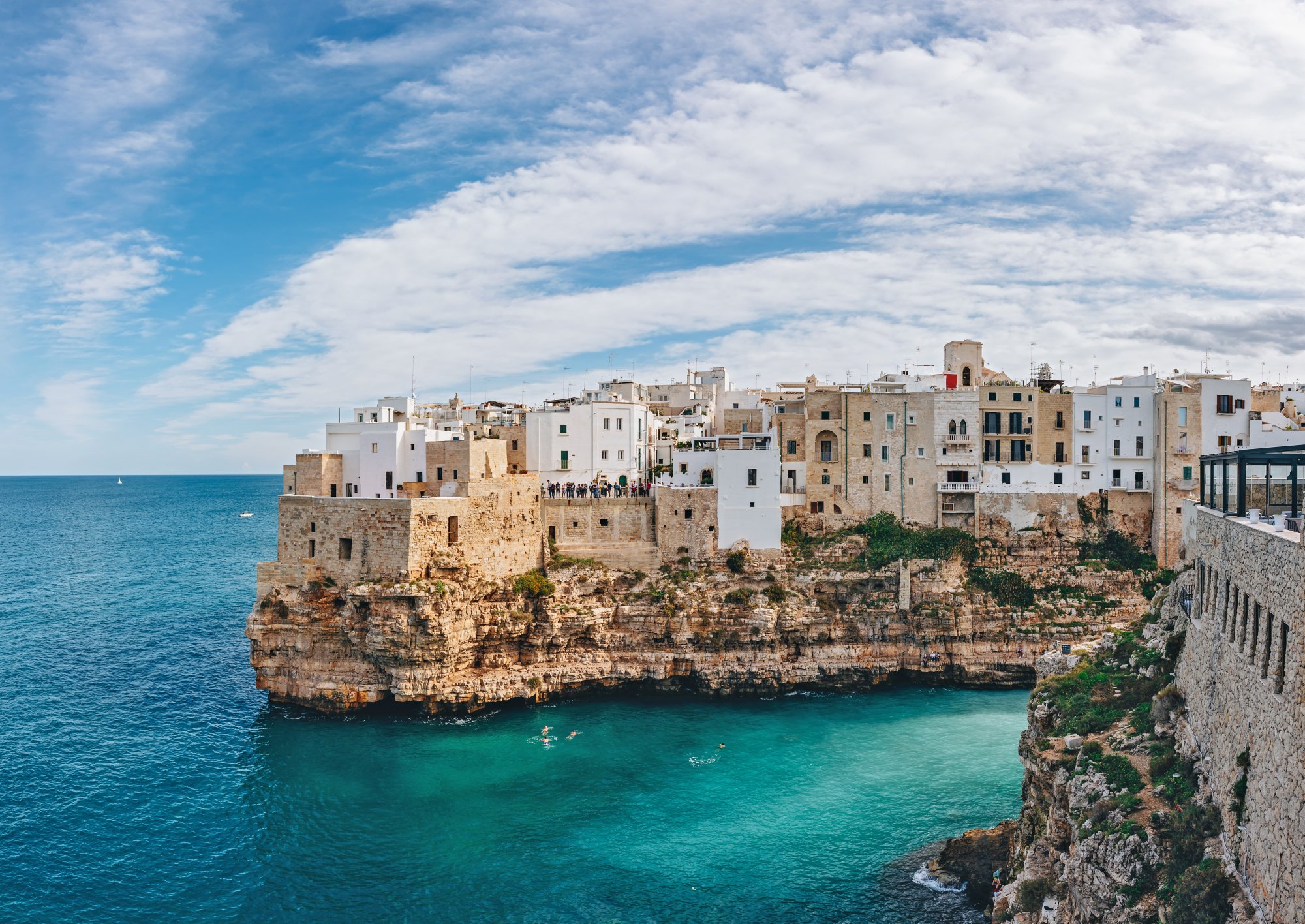 Küstenstadt Bari mit weißen und beigen Gebäuden auf Felsen über türkisfarbenem Meer und Schwimmern nahe der Klippen.