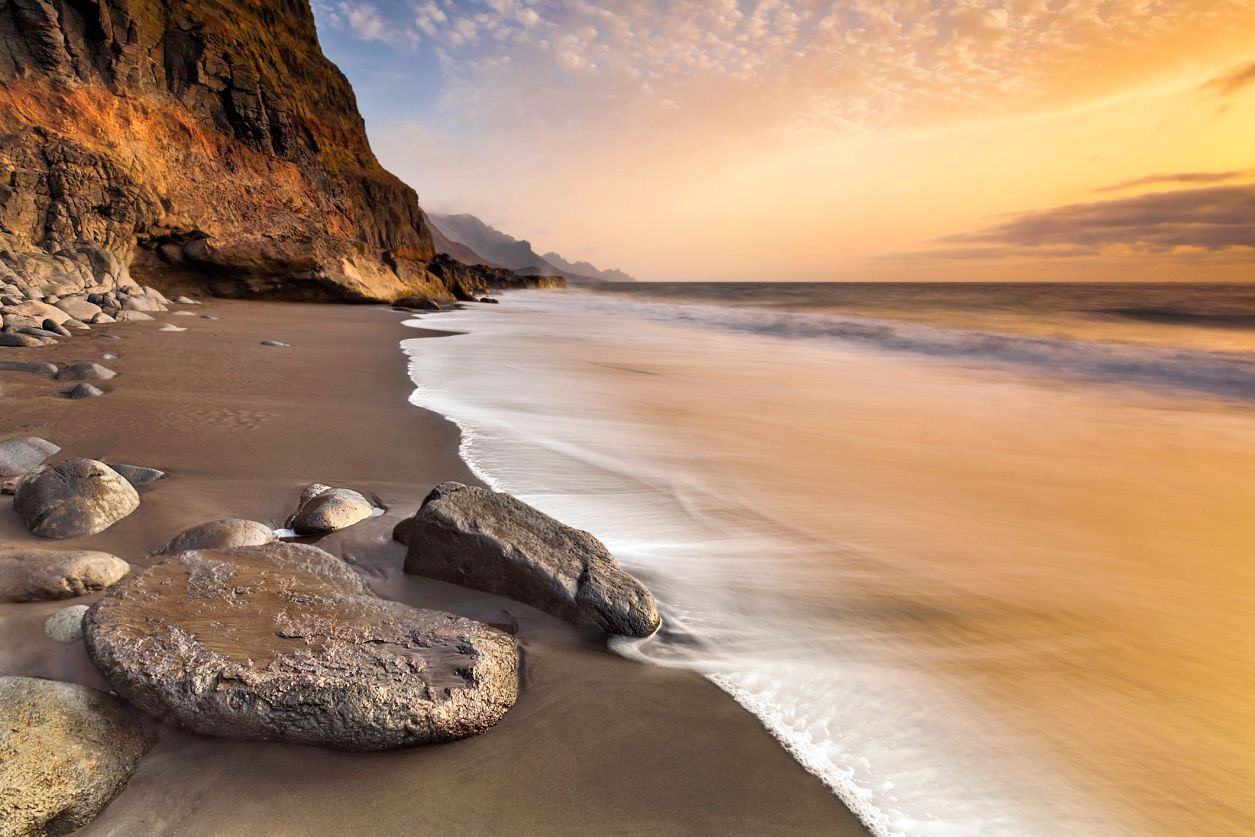 Strand mit Felsen, sanften Wellen und goldenem Himmel bei Sonnenauf- oder -untergang.