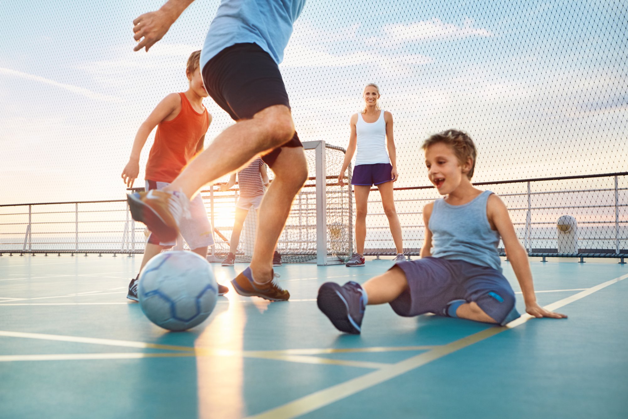 Eine Familie mit Mutter, Vater und 3 Kinder spielt Fußball auf dem Sportdeck. Die Sonne geht im Hintergrund unter.