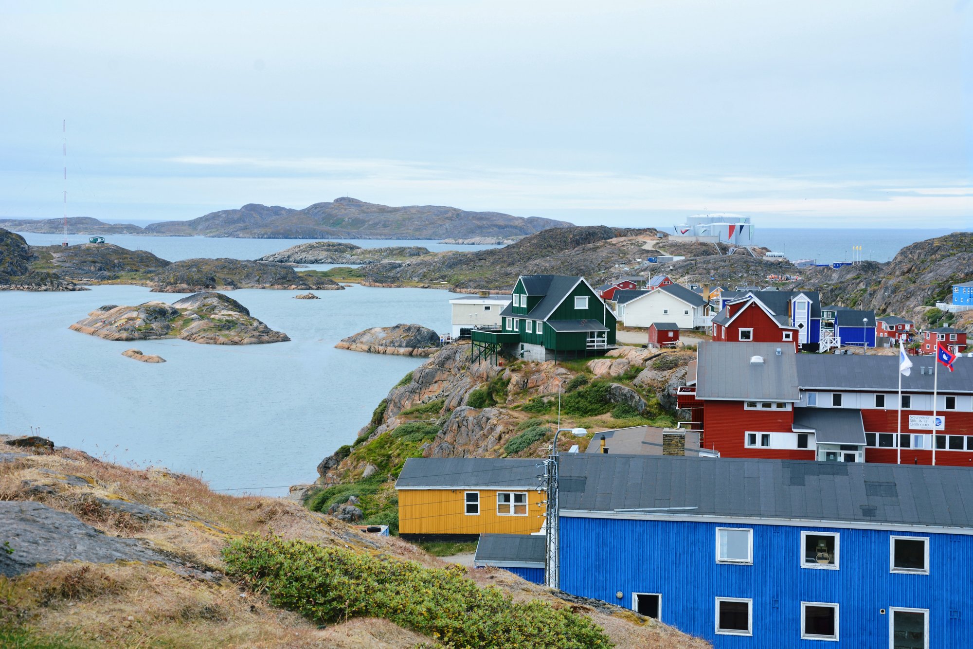Bunte Häuser auf felsigem Gelände in Sisimiut, Grönland, mit Blick auf eine ruhige Bucht unter bewölktem Himmel.