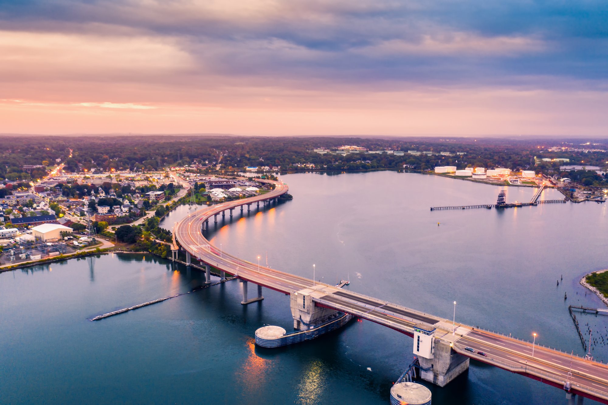 Eine große Brücke mit mehreren Fahrspuren überspannt ein Gewässer und verbindet städtische und industrielle Bereiche bei leicht bewölktem Himmel.