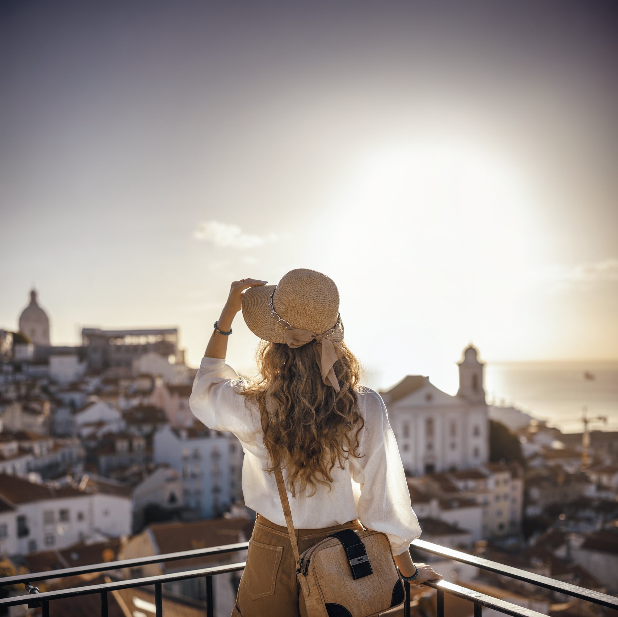 Blonde woman standing on the balcony and looking at coast view of the southern european city with sea during the sunset, wearing hat, cork bag, safari shorts and white shirt