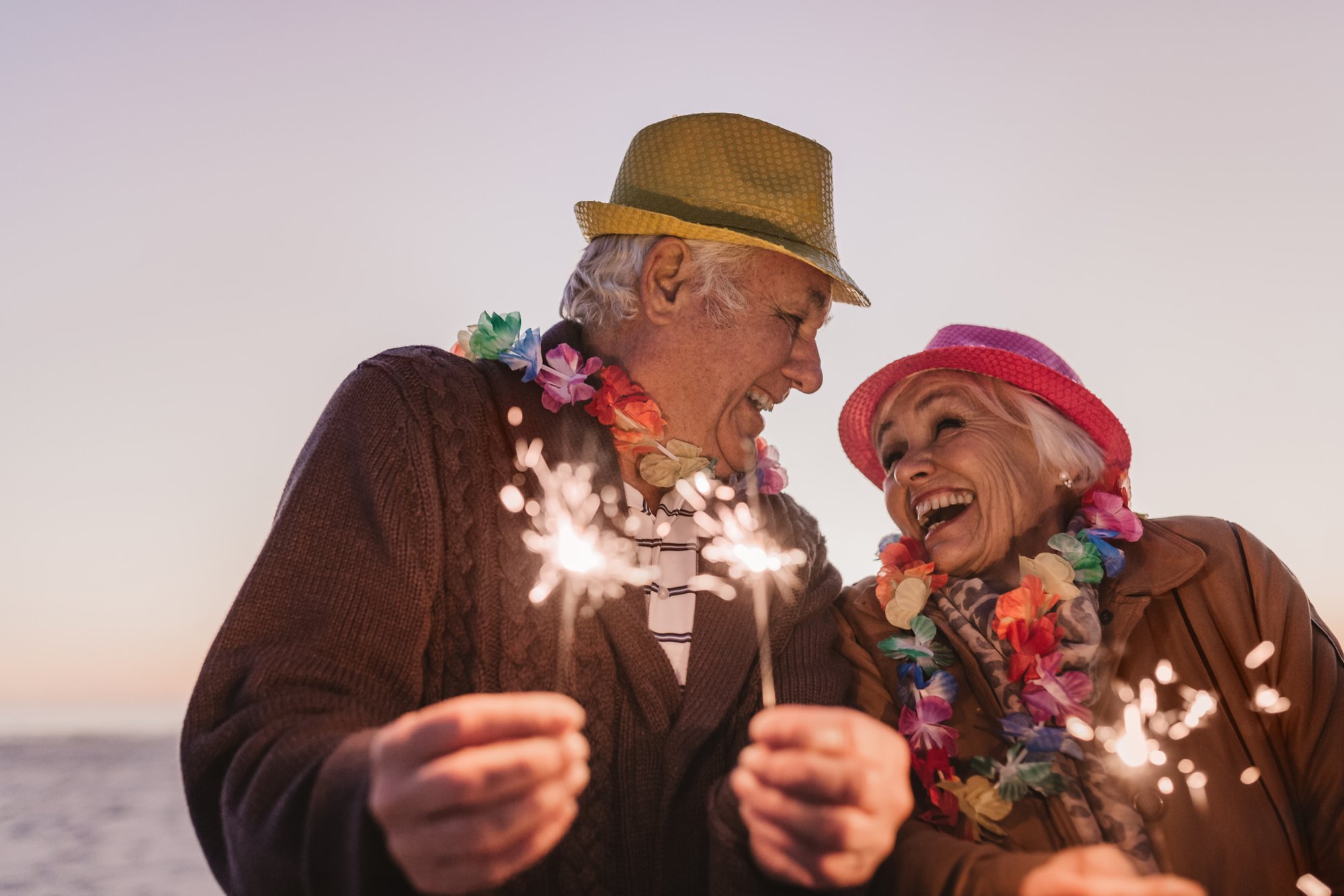 Laughing senior couple dressed in party hats and holding sparklers while celebrating New Year's Eve together at the beach at sunset