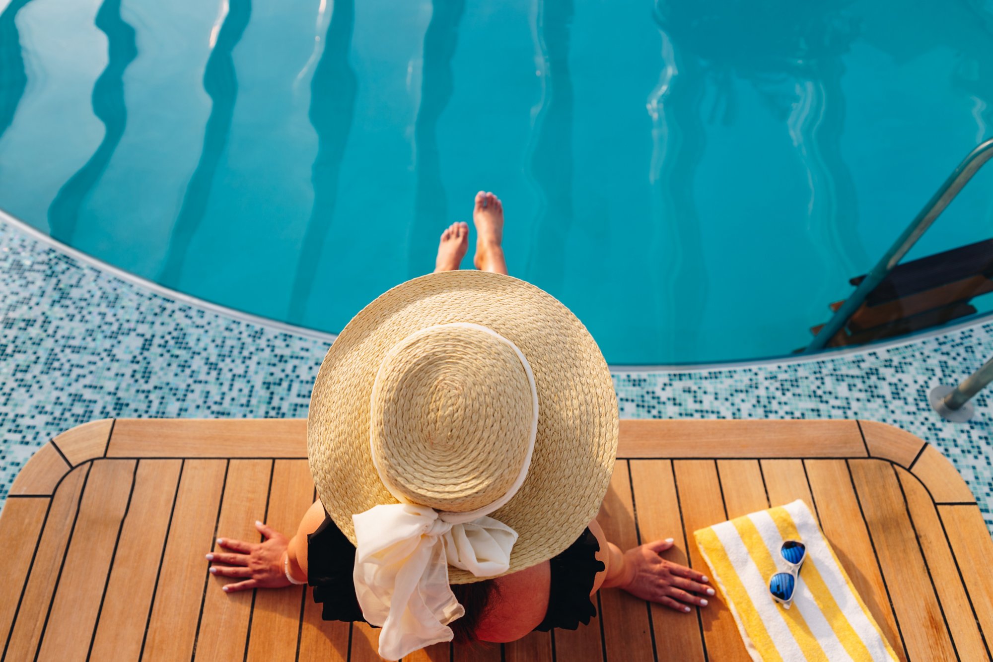 Eine Frau mit einem Sonnenhut sitzt am Pool und lässt die Füße ins Wasser hängen, man sieht sie von oben