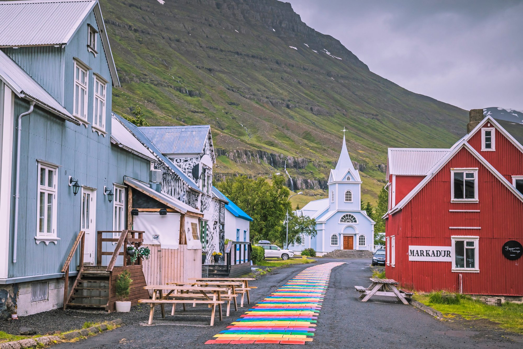 Eine blaue Kirche in Seydisfjördur mit einem bunten Gehweg im Vordergrund und einem großen Berg im Hintergrund..