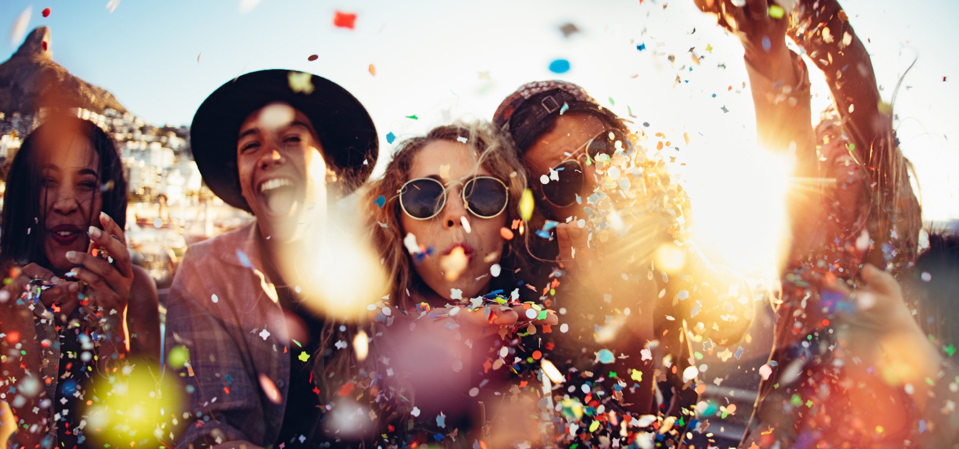 Group of teenager hipster friends partying by blowing and throwing colorful confetti from hands with sunset sun flare