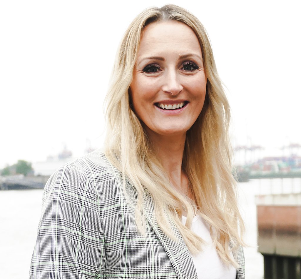 Frau mit langen hellblonden Haaren, geschminkten Augen und einem leichten Lächeln. Sie trägt einen hellgrau weiß karrieten Blazer.