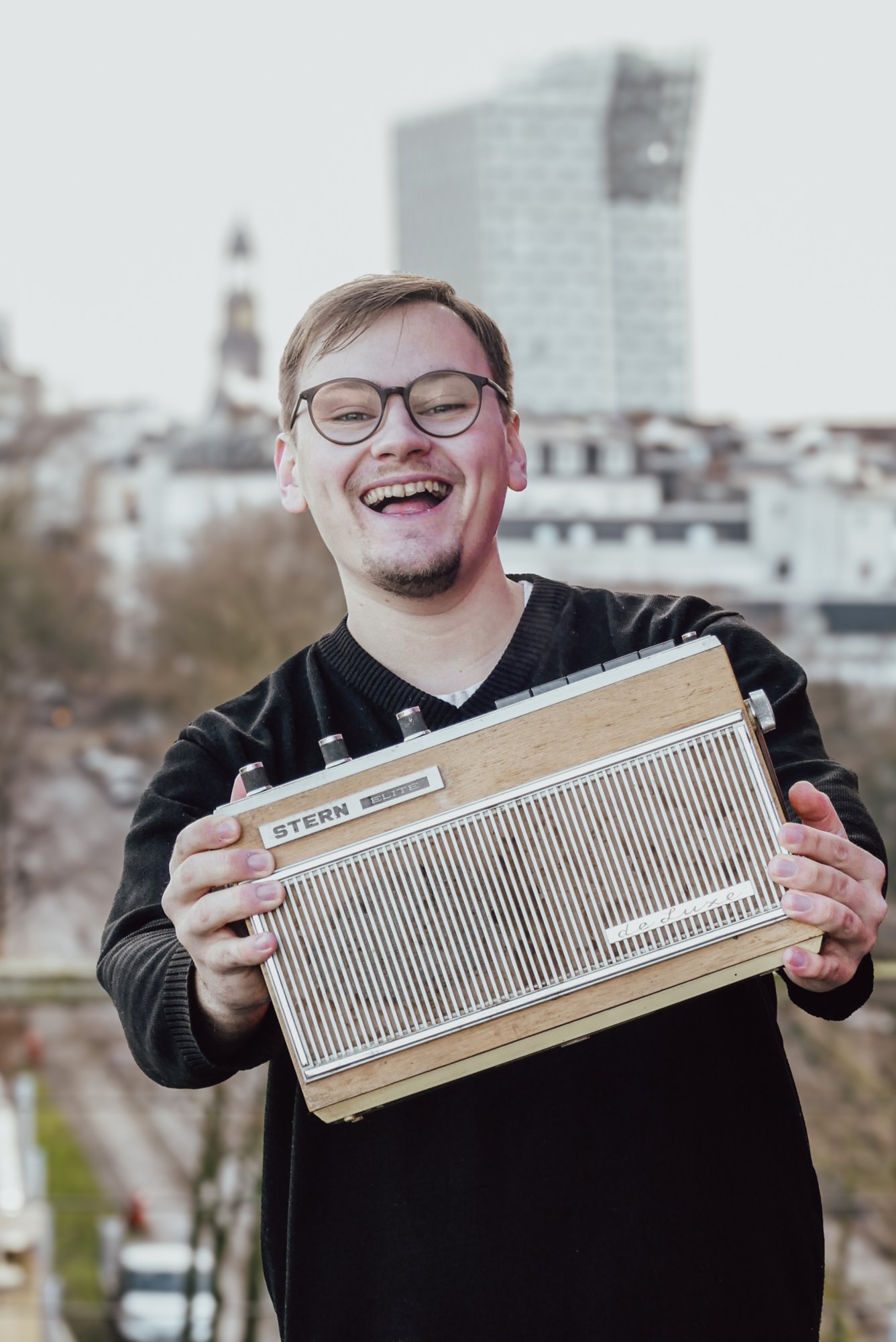 Junge Mann mit Brille und Kinnbart, trägt einen schwarzen V-Ausschnitt Pullover und hält ein altes Radio in die Kamera. Er lacht dabei.