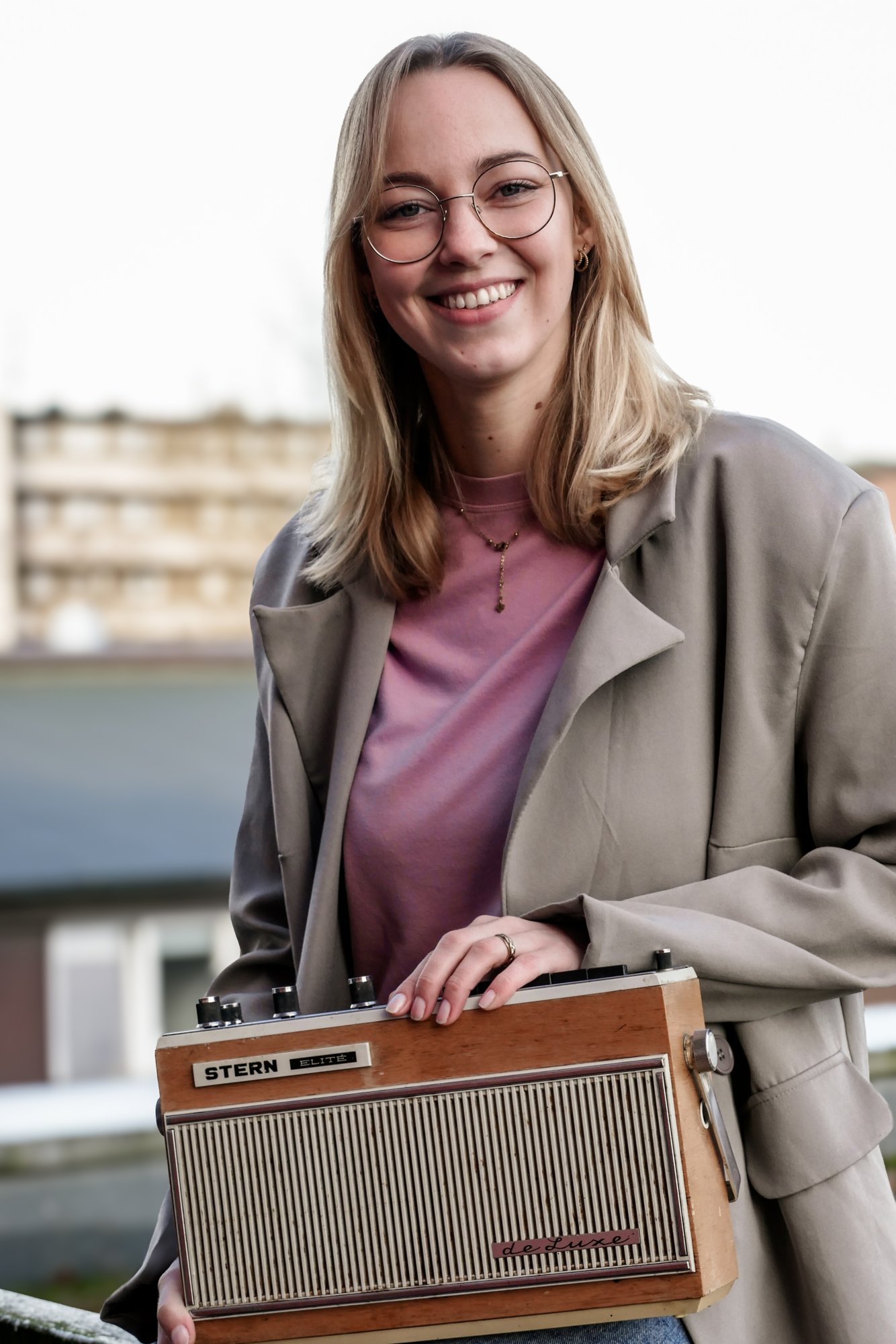 Junge Frau mit blonden schulterlangen Haare und Brille. Sie trägt ein fliederfarbenes T-Shirt, darüber eine beige Jacke. Sie lächelt in die Kamera und hält ein altes Radio in den Händen.