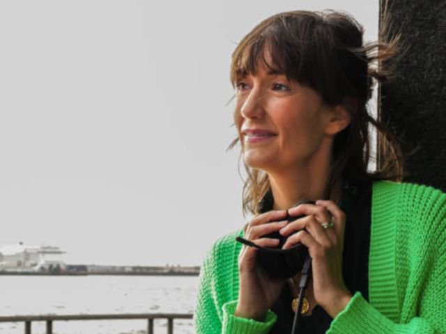 Frau mit langen braunen Haaren und leichtem Lächeln. Sie hält ihre Hände auf Schlüsselbeinhöhe und hält ein Headset in den Händen. Sie trägt ein neongrünen Strickjacke und eine schwarze Bluse.