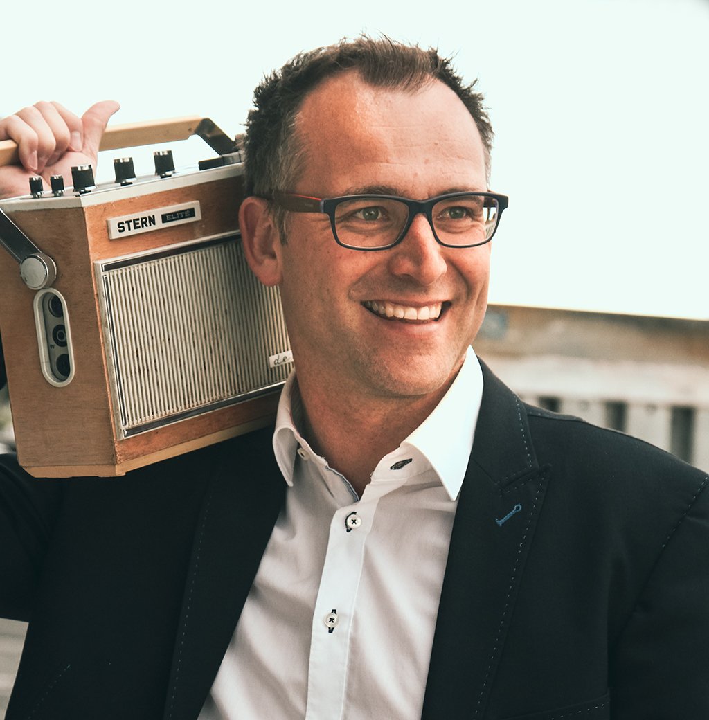 Mann mit schwarzer Brille und einem Breiten lächeln mit Zähnen. Er hat schwarz graue Haare und trägt eine schwarze Anzugjacke und darunter ein weißes Hemd.