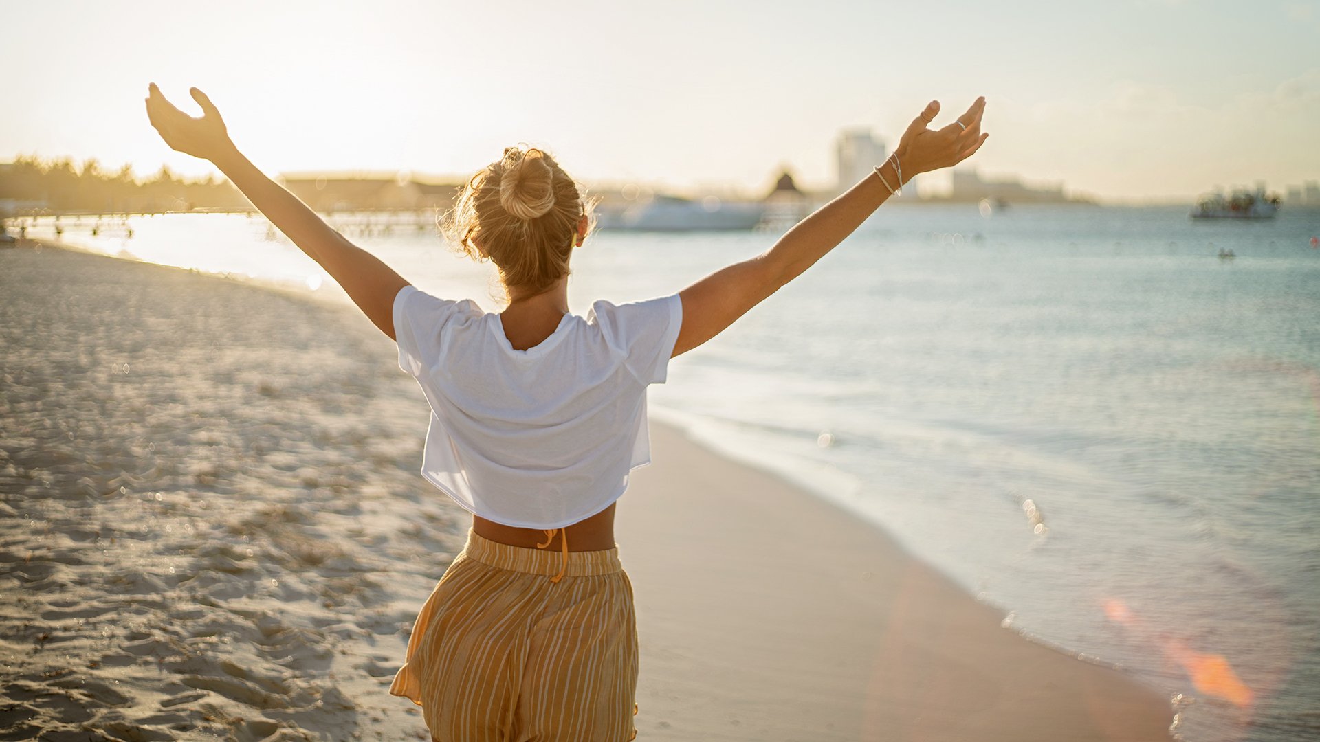 Cheerful young woman embracing nature at sunset; female standing on beach arms outstretched, Cancun, Mexico